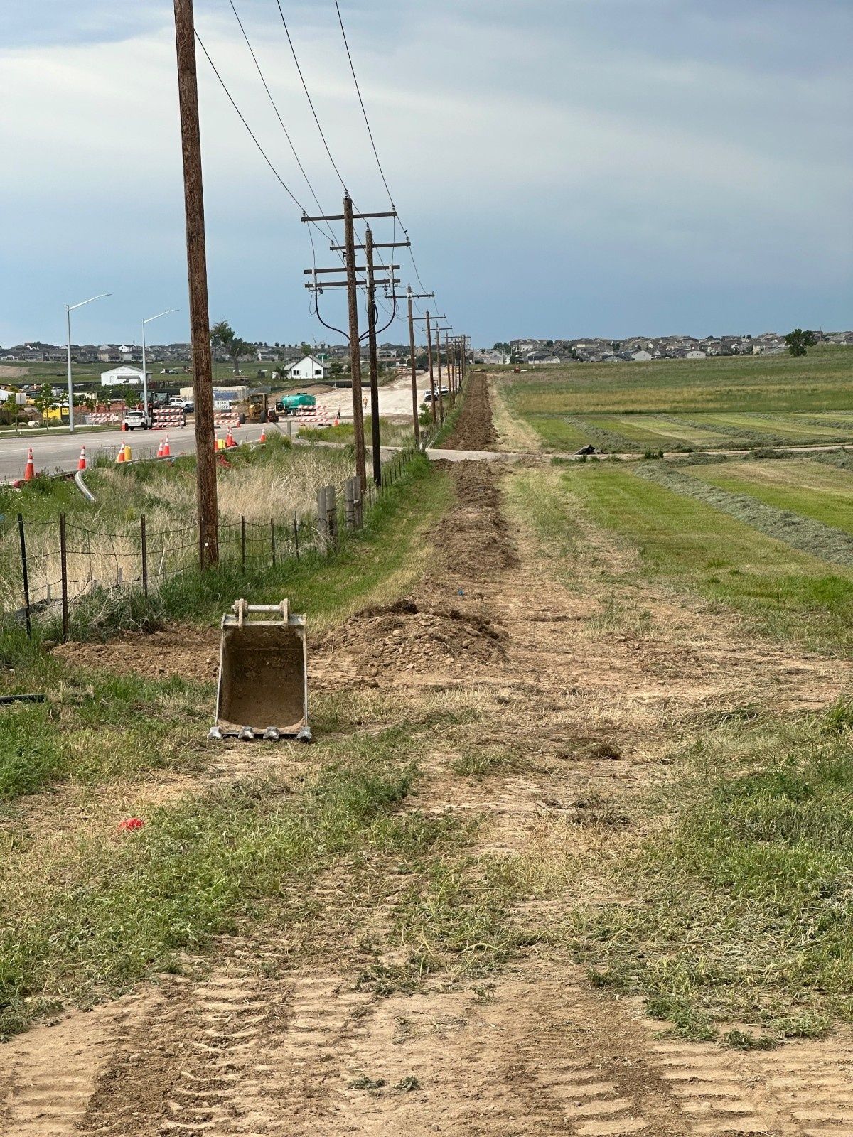 A dirt path runs alongside utility poles and a grassy field, with an excavator bucket resting in the foreground.