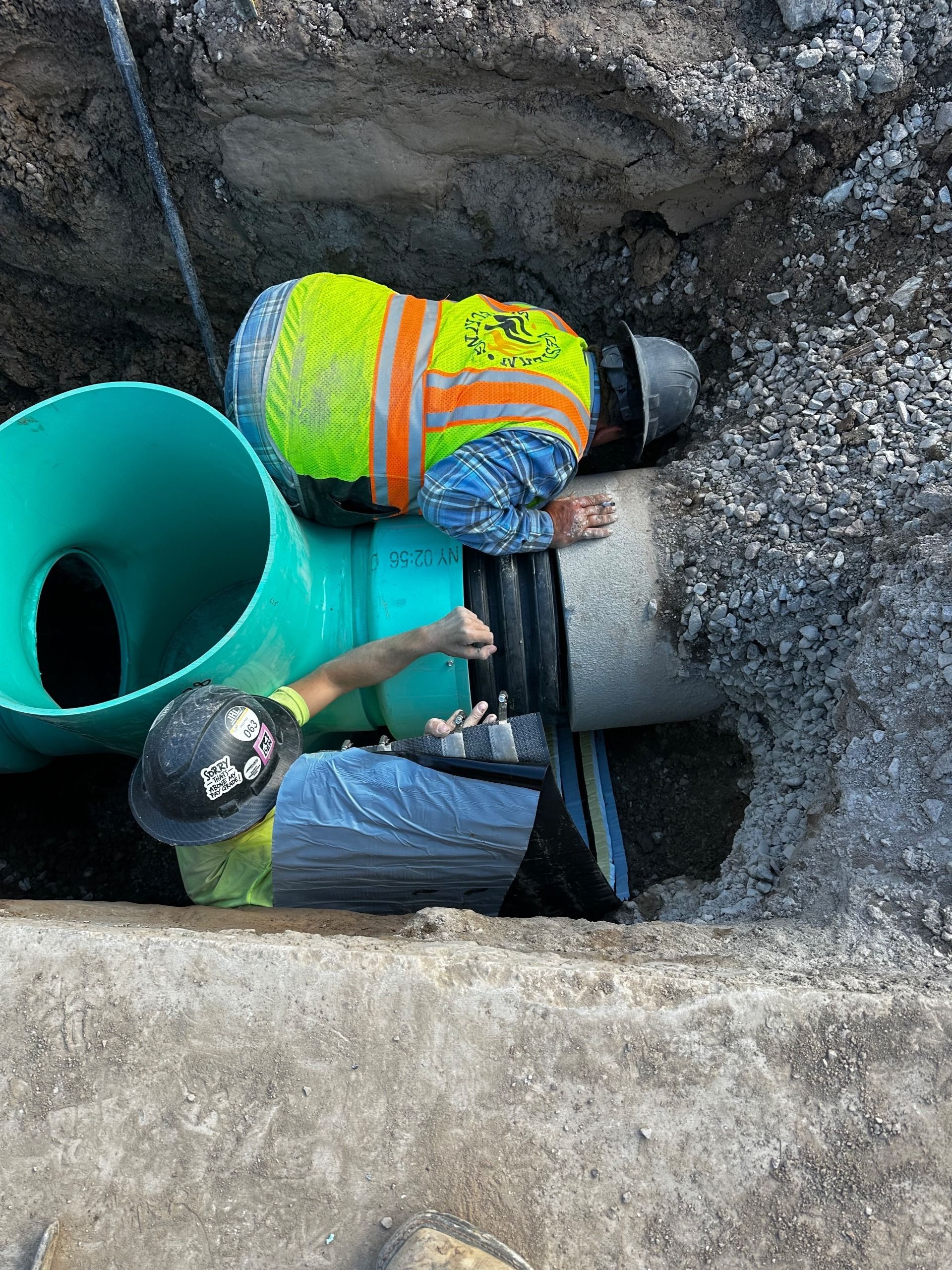 Two construction workers in high-visibility vests and hard hats install a large green pipe in an earthen trench.