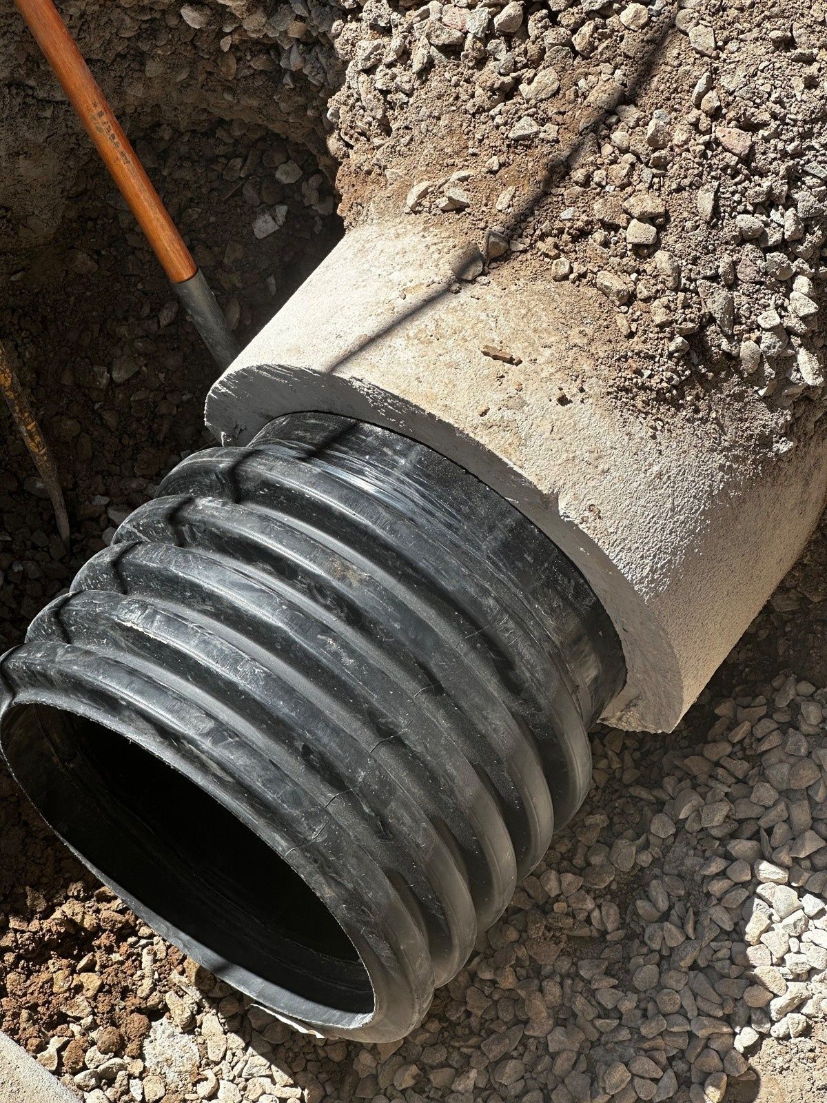 A black corrugated plastic pipe extending from a thick concrete pipe in a dirt trench, with a shovel handle visible.