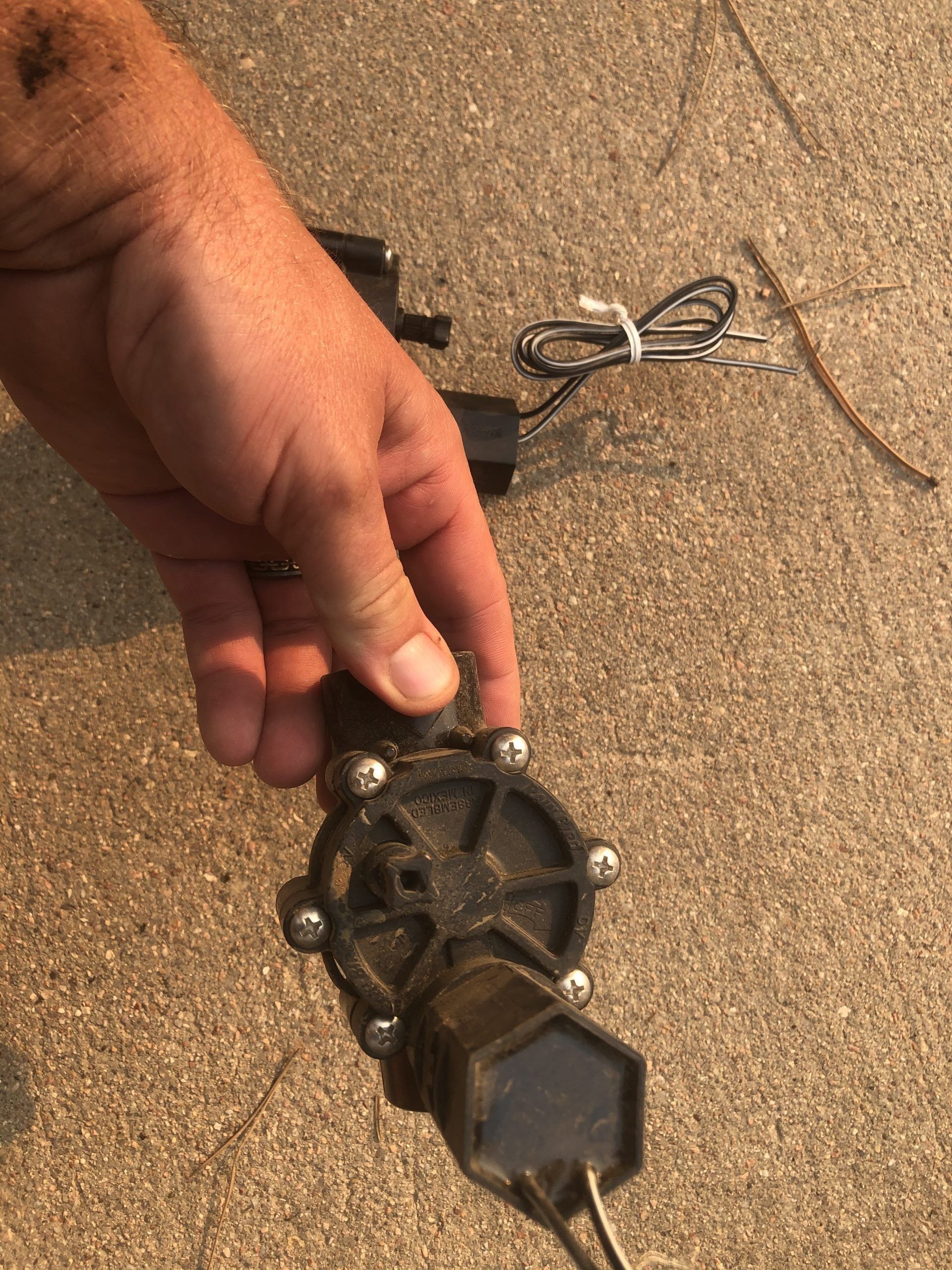 A hand holding a dirty black irrigation sprinkler valve with exposed electrical wires against a sandy background.
