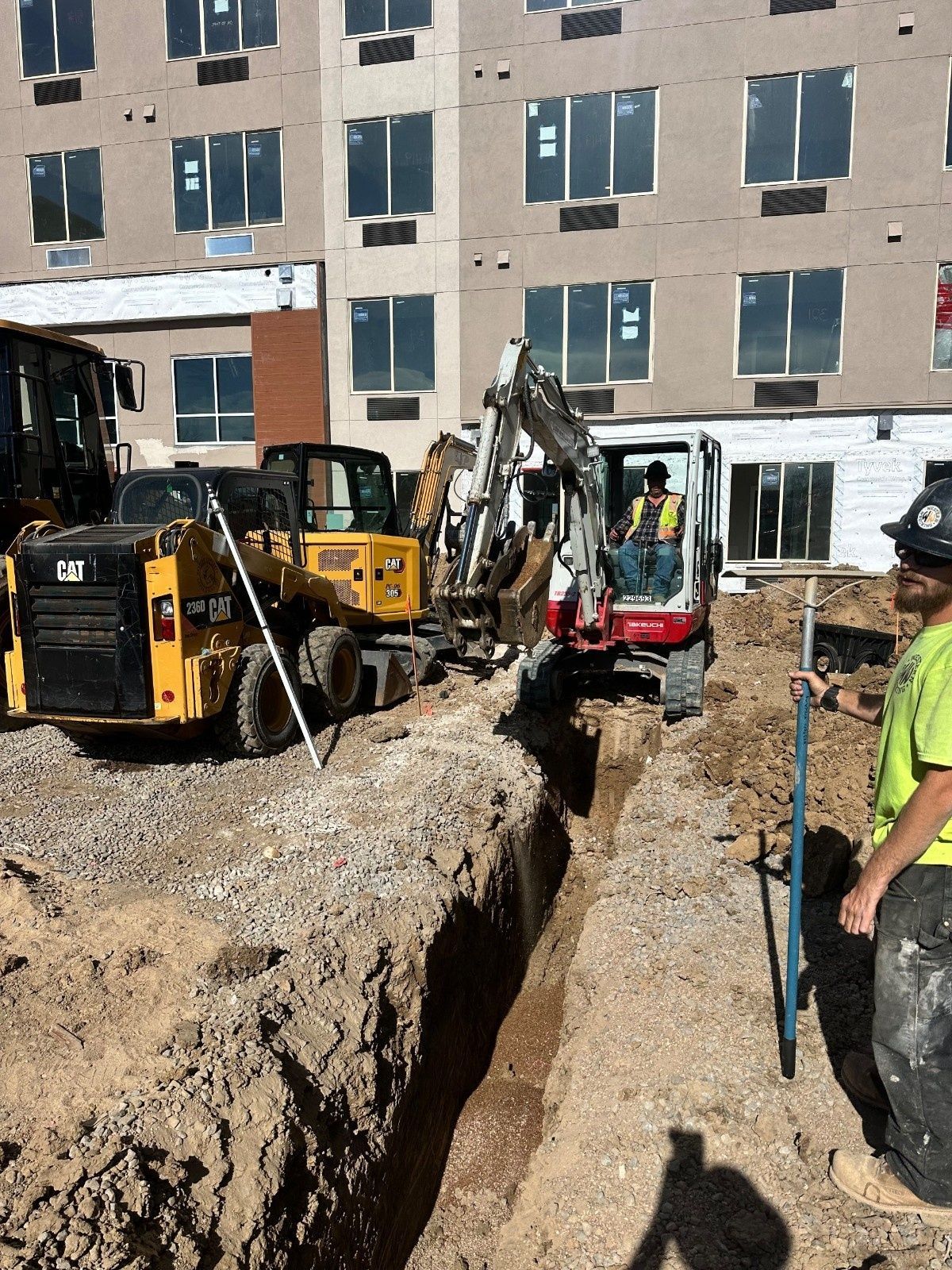 Construction workers use a yellow skid steer and a small excavator to dig a trench in front of a multi-story building.