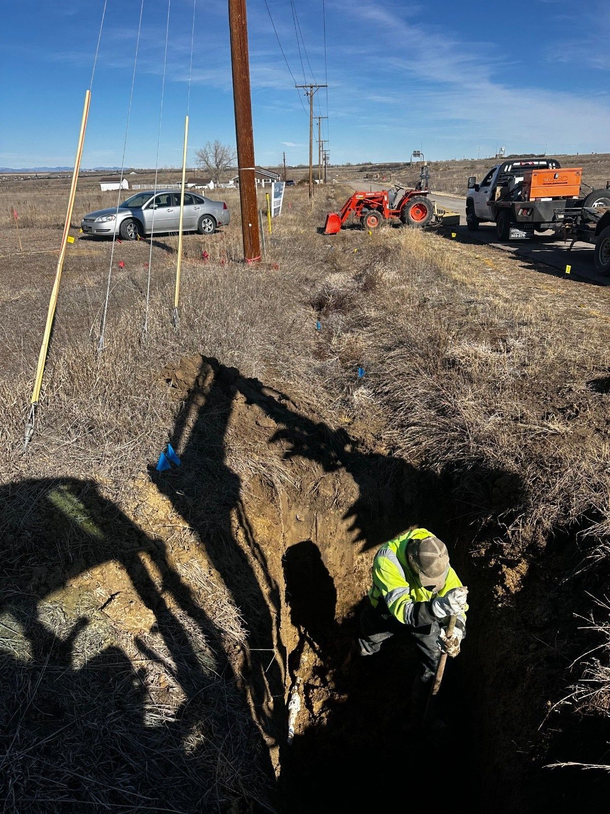 A worker in a high-visibility jacket repairs a utility line in an open trench near a parked truck and tractor.