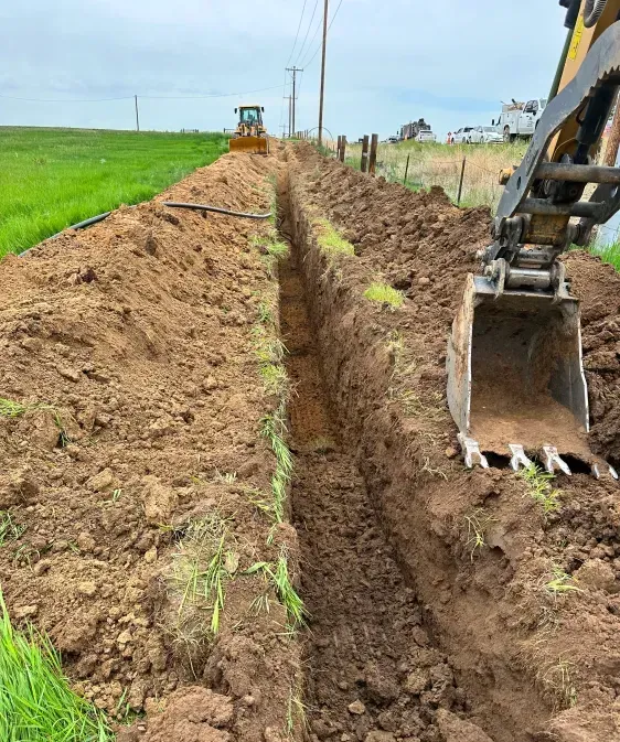 An excavator digging a long, deep trench through an open field with a bulldozer visible in the background.