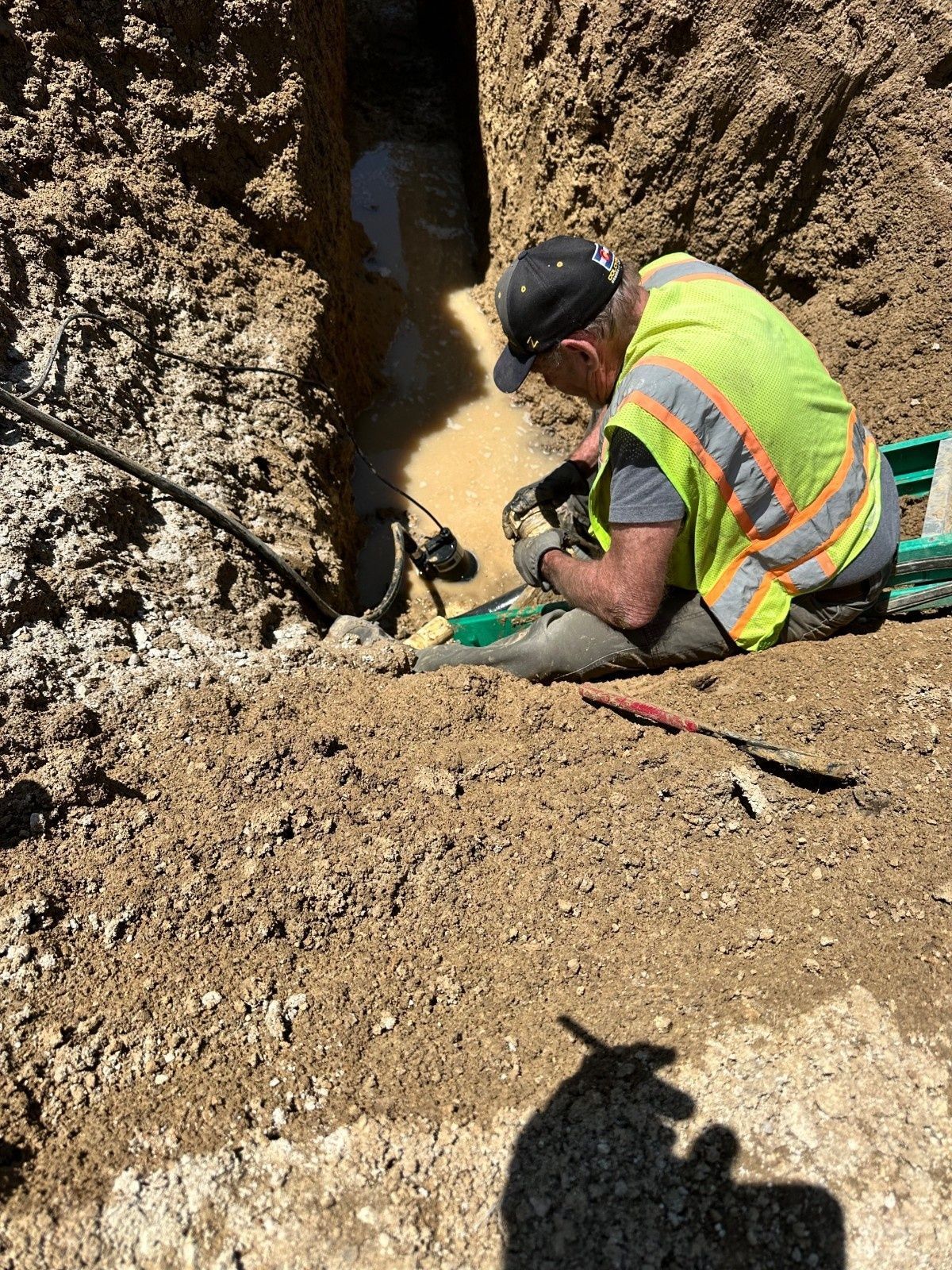 A worker in a high-visibility vest kneels at the edge of a muddy trench, working on utility pipes.