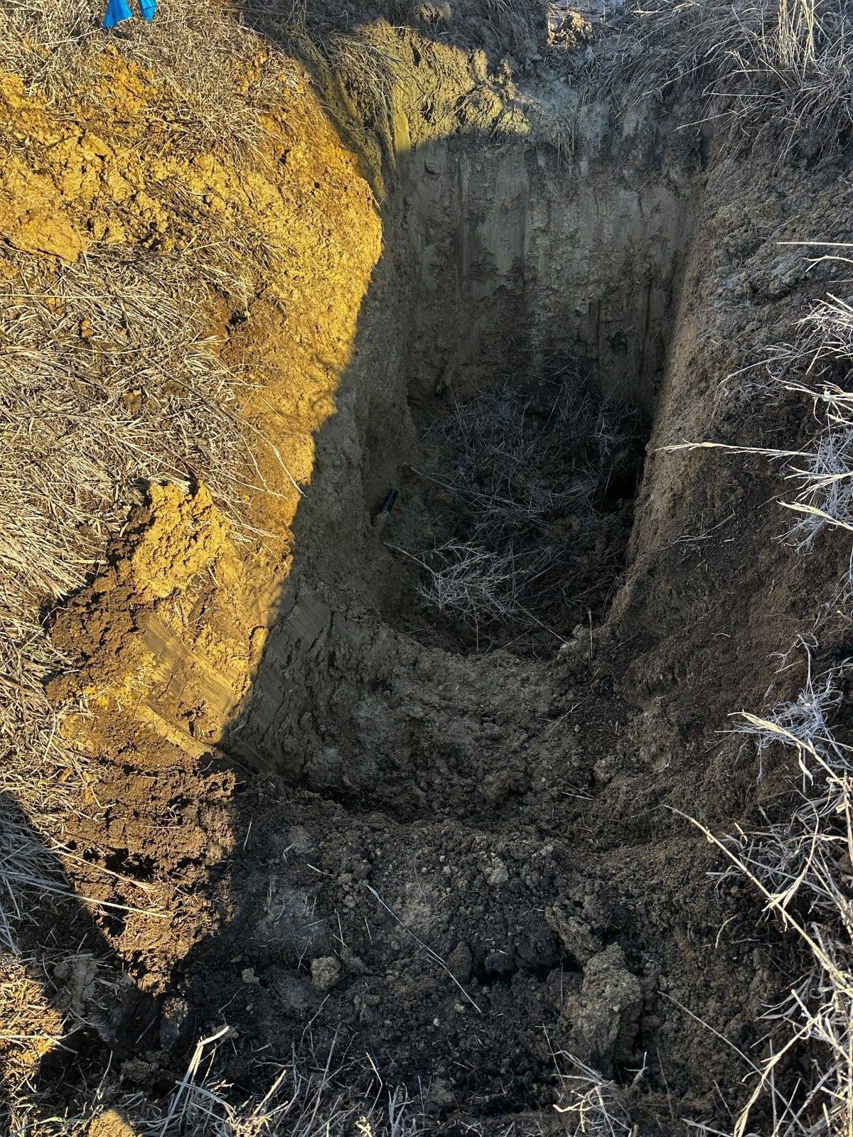 A rectangular trench dug into the ground, revealing layers of dark topsoil over a dense, lighter-colored subsoil.