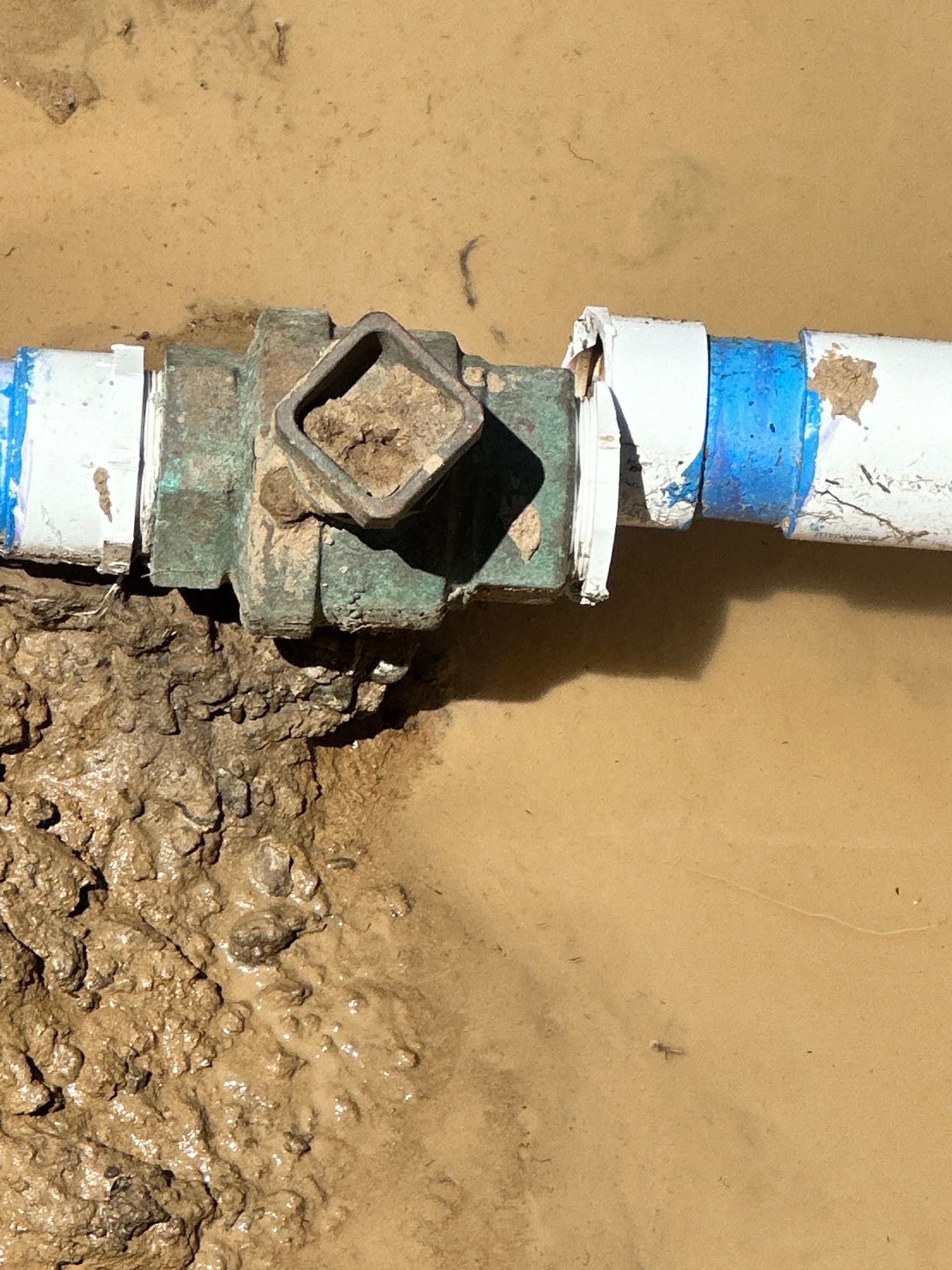 A close-up of a damaged, blue-banded white pipe connected to a green metal valve sitting in muddy ground.