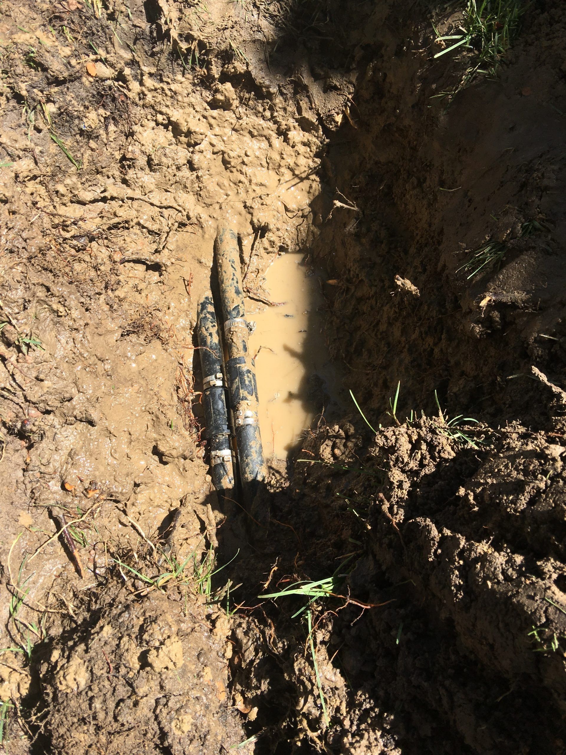 Two dark, cylindrical pipes run parallel through a muddy, dirt-filled trench with pooled water.