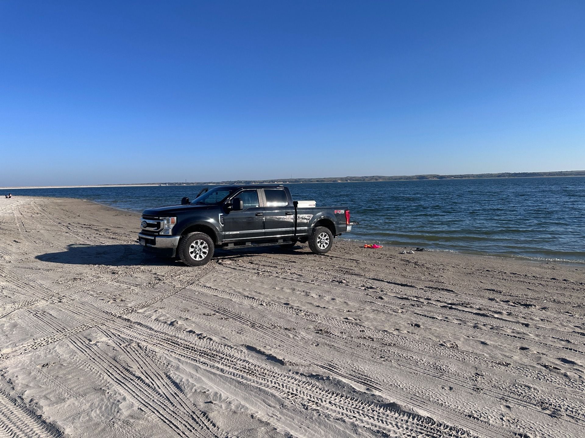 A dark pickup truck parked on a sandy beach beside a calm, blue body of water under a clear, bright sky.