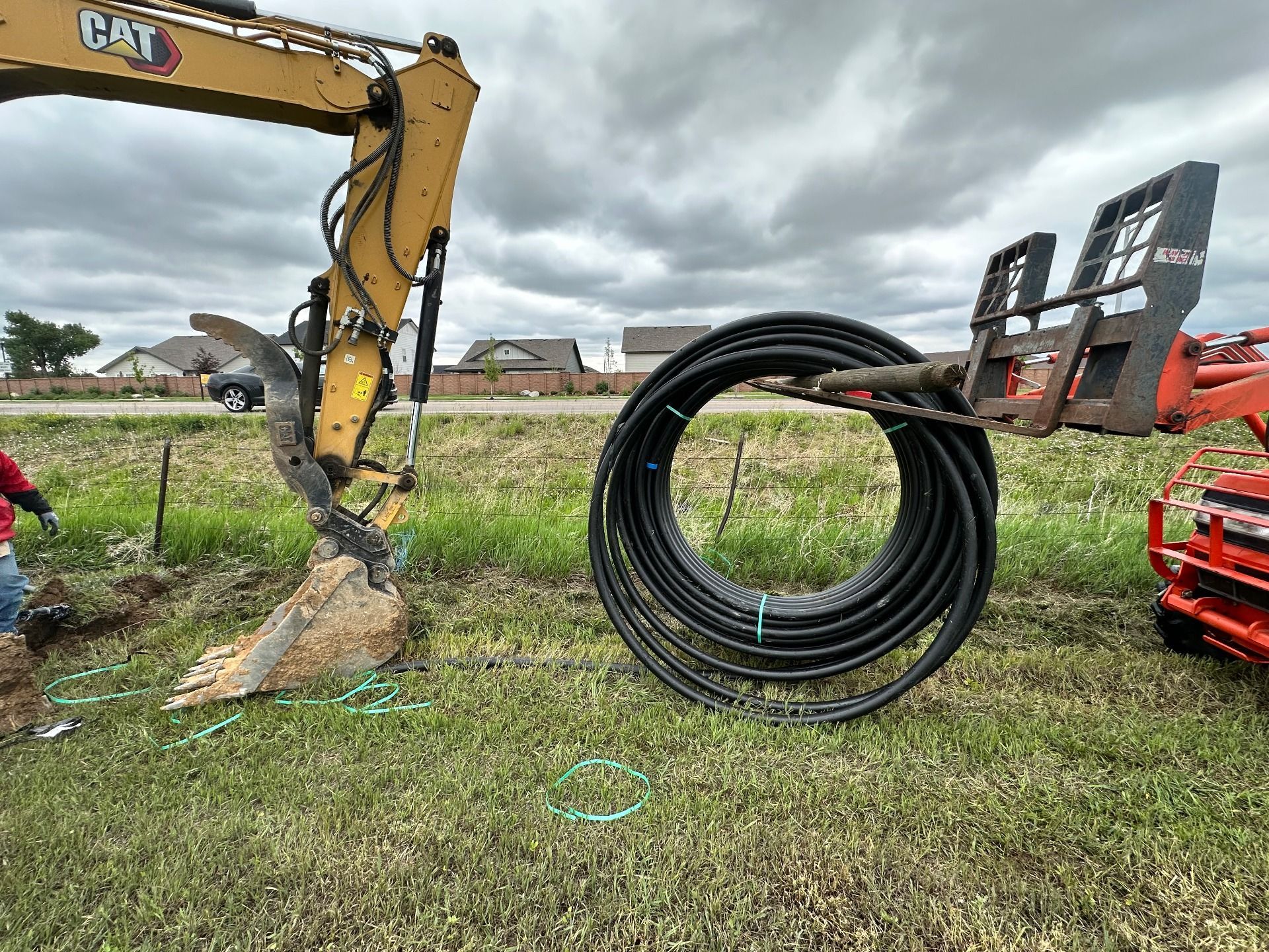 An excavator and a skid steer with forks holding a large coil of black plastic pipe on a grassy construction site.