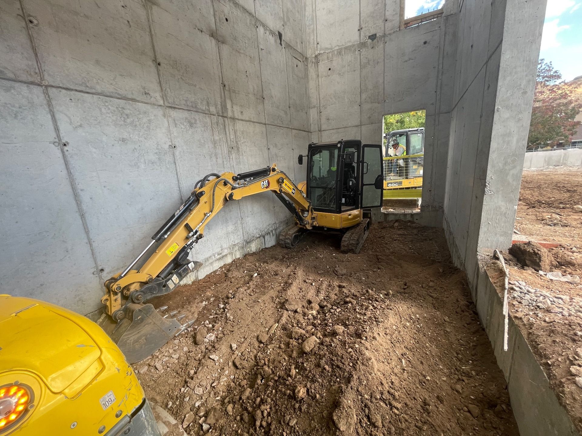 A yellow compact excavator maneuvers inside an unfinished concrete room, with a view of a dirt lot visible through a gap.