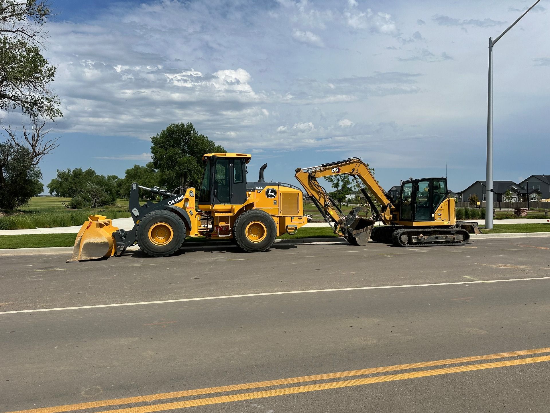 A yellow front loader and a yellow excavator parked side-by-side on the side of a road against a blue, cloudy sky.