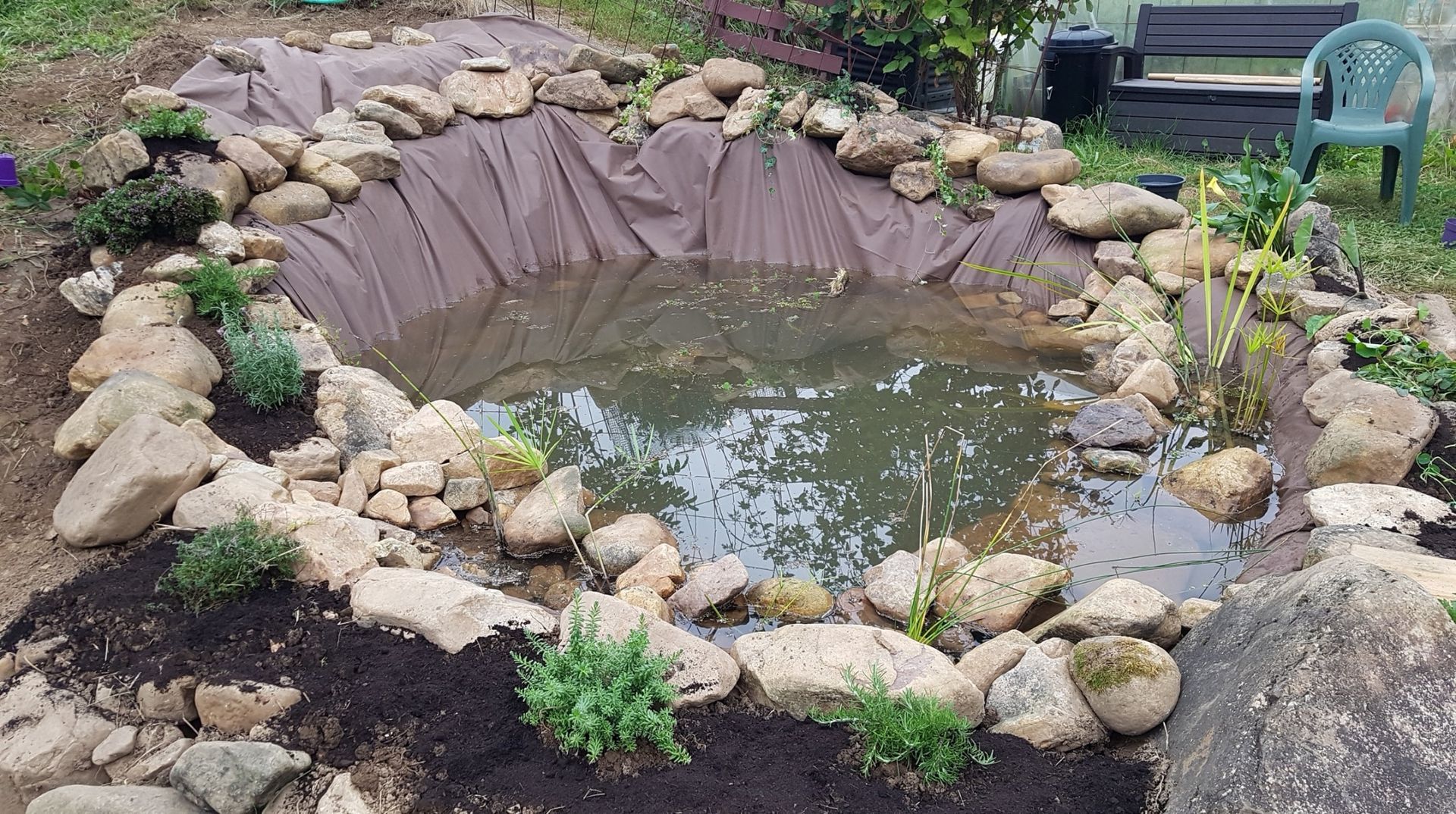 A newly constructed garden pond lined with dark fabric, bordered by rocks and small plants, with a bench in the background.