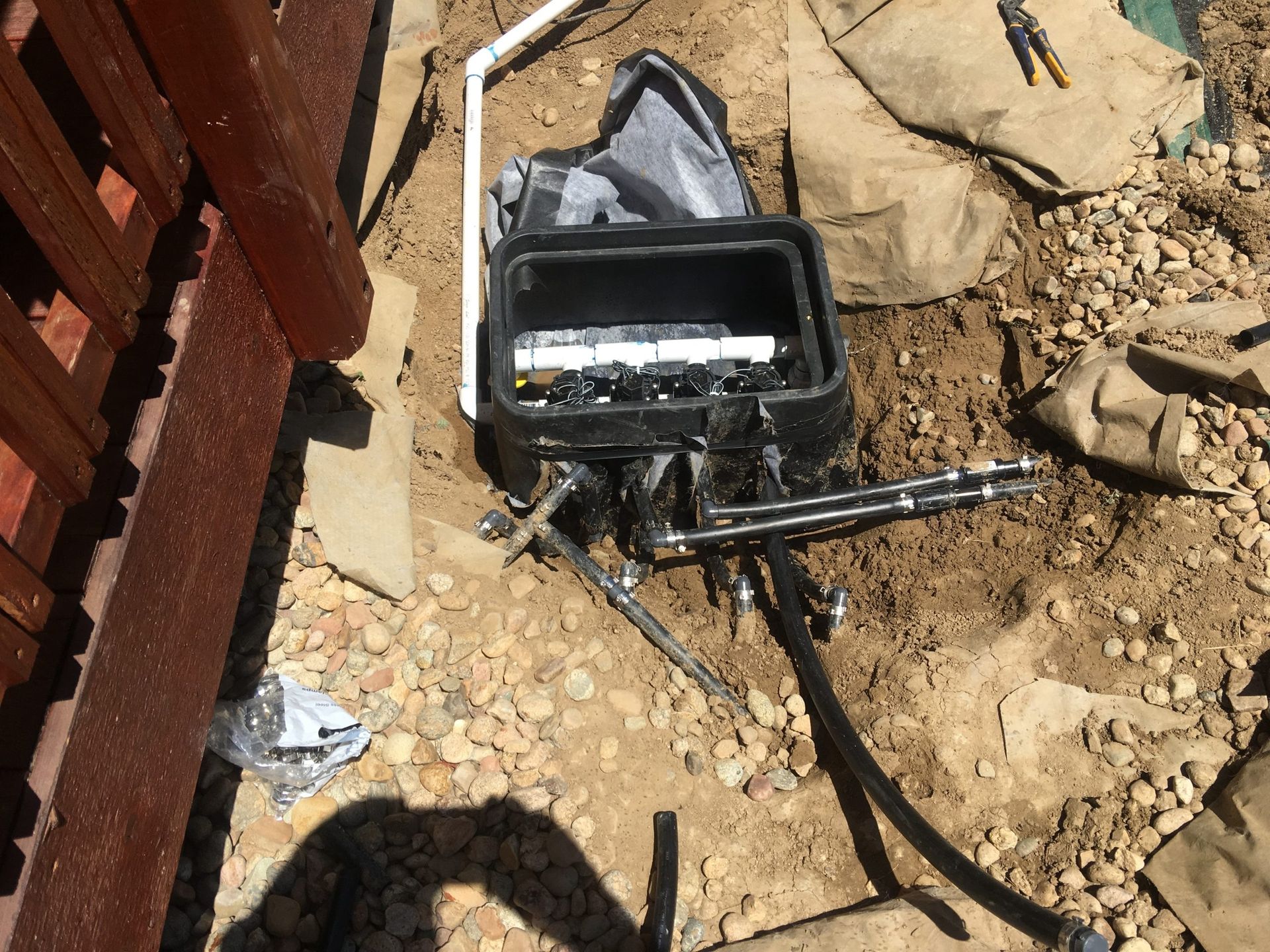 An irrigation valve box with exposed pipes and black tubing, installed in a dirt and gravel area beside a wooden fence.