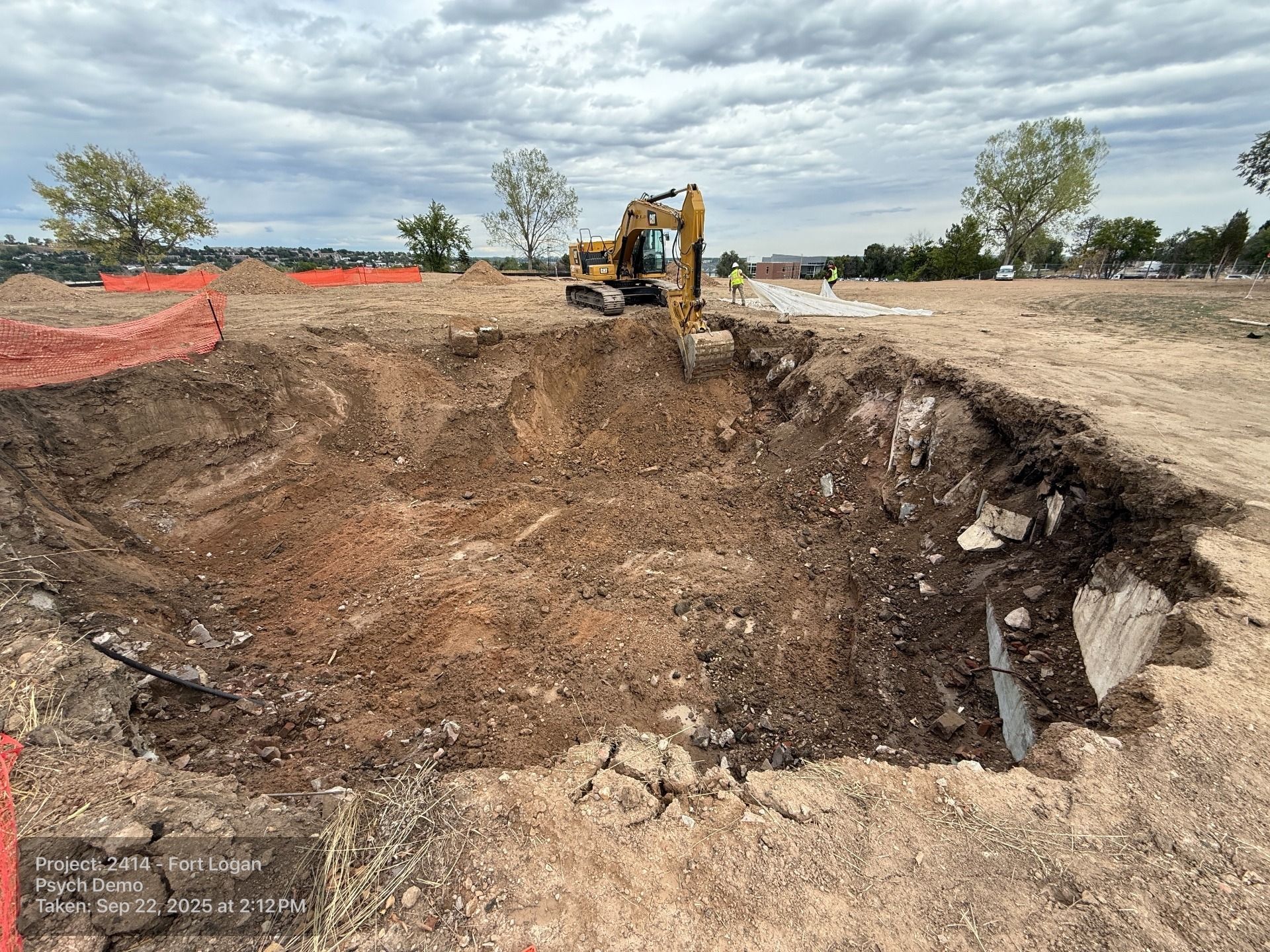 A yellow excavator digs into a large, rectangular pit in a dirt construction site under a cloudy sky.