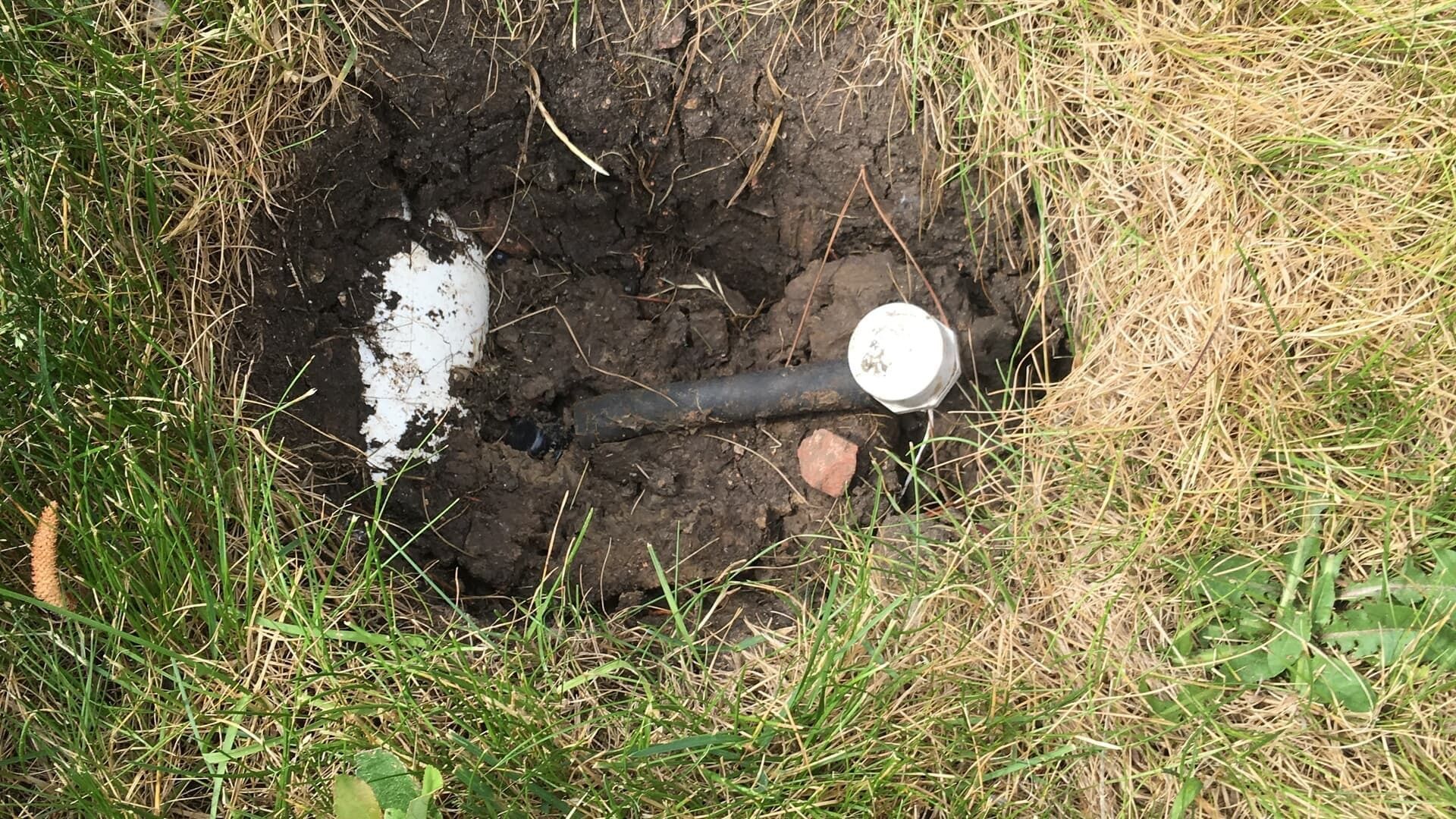 A dark circular hole in a lawn revealing a white PVC pipe section and a black irrigation pipe with a white cap.