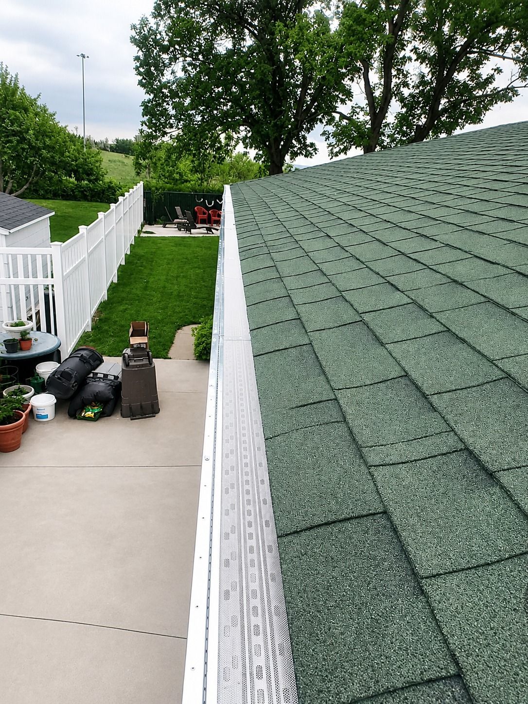 White rooftop deck beside a green shingle roof, with potted plants and tools along a narrow walkway.
