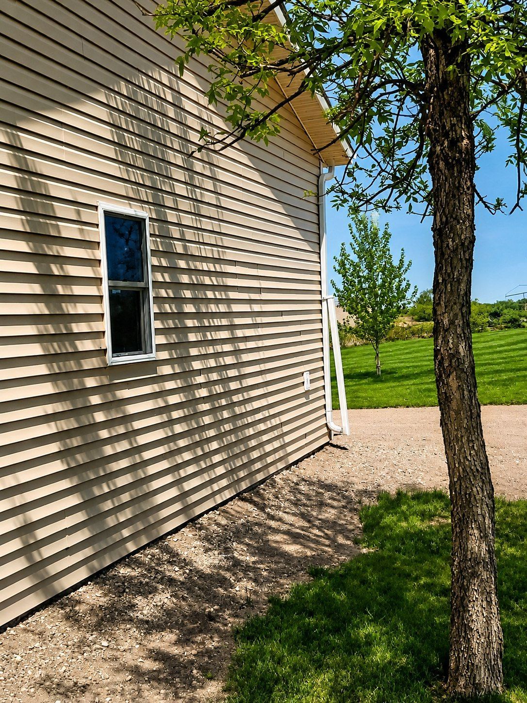 Side of a beige house with a small window, tree trunk, gravel path, and sunny green lawn.