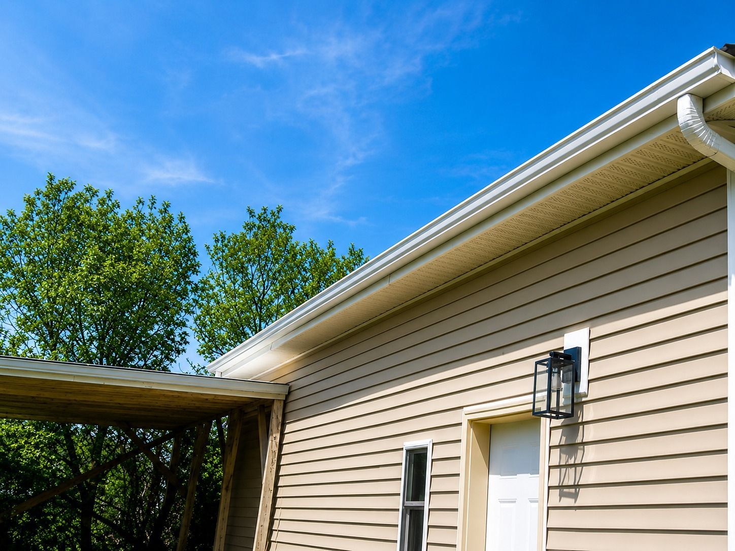 Beige house exterior with white door, gutter, and a blue sky above trees