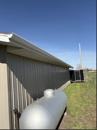 Metal storage tank beside a tan building under a clear blue sky, with grass and a distant fence line.