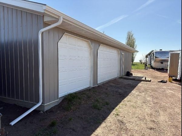 Row of white garage doors on a tan metal building beside a dirt driveway under a clear blue sky