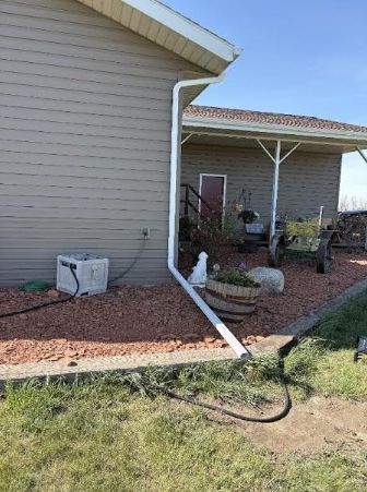 Backyard side of a beige house with white downspout, covered patio, rocks, and garden hose on grass