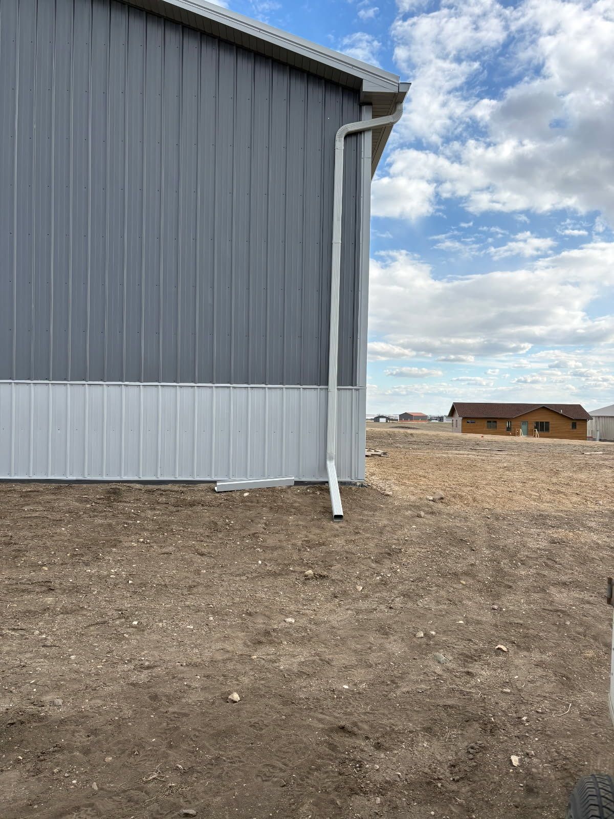 Gray metal building corner beside a bare dirt lot under a cloudy sky with a small shed in the distance