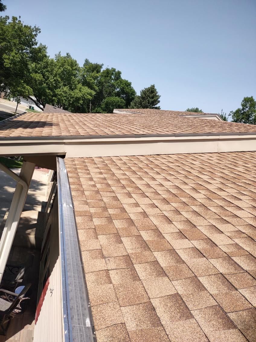 Roof with tan shingles viewed from above beside a tree-lined backyard under a clear blue sky