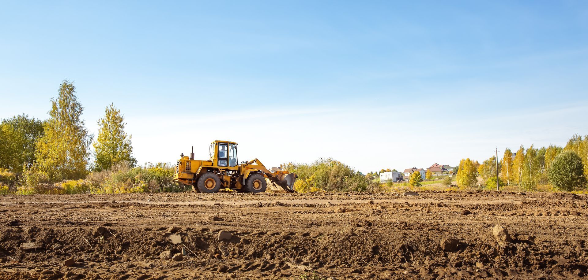 A yellow bulldozer working in a brown field under a blue sky. Trees in the background.