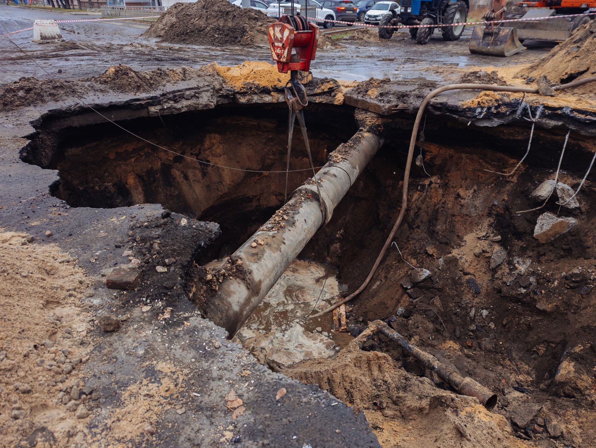 Large hole in road revealing damaged pipe, crane above, workers in background.