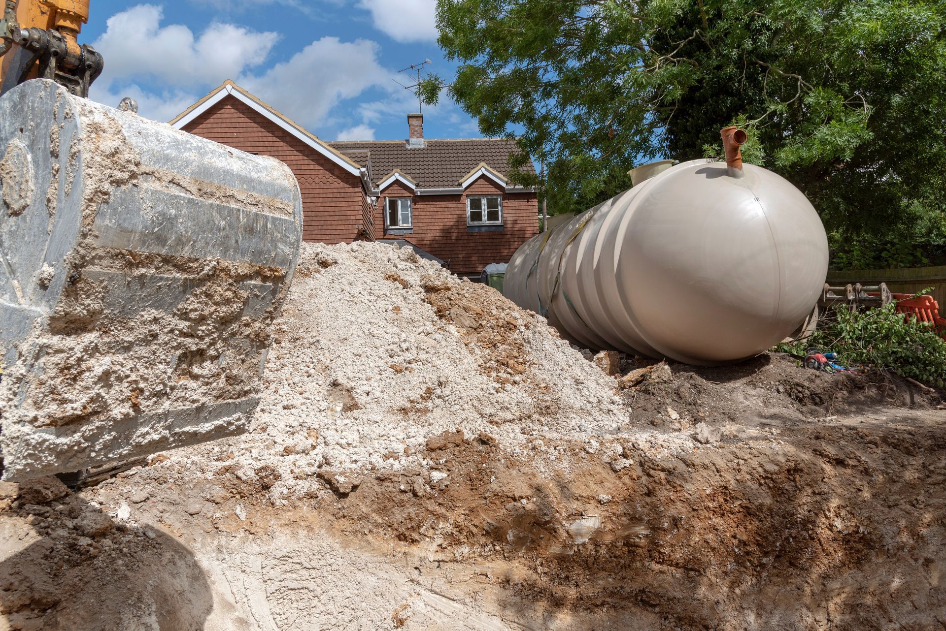 Excavator next to a buried tank in a construction site, soil mound, houses in background.