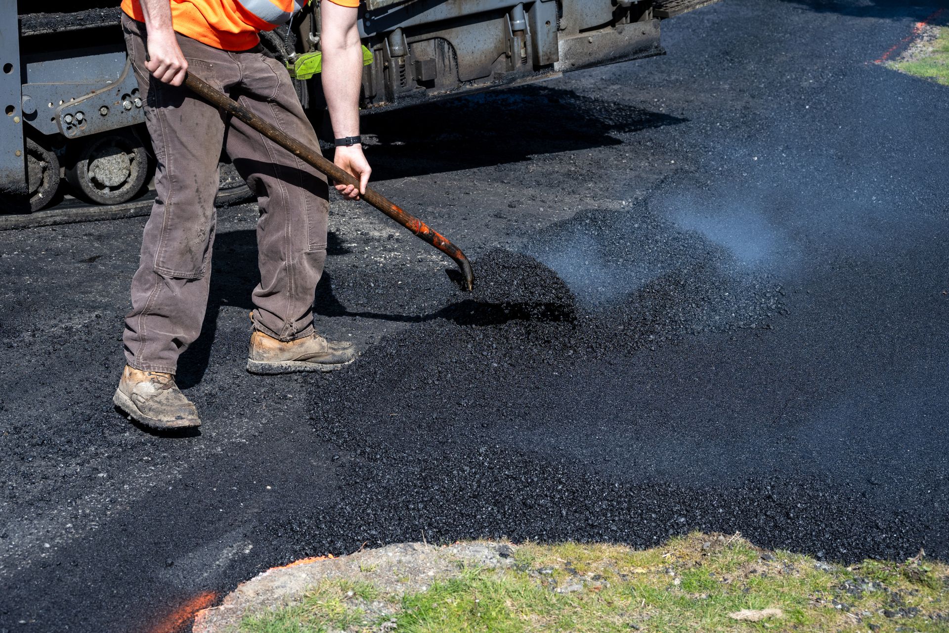 Worker using a rake to spread steaming asphalt on a road; asphalt paving machine in background.