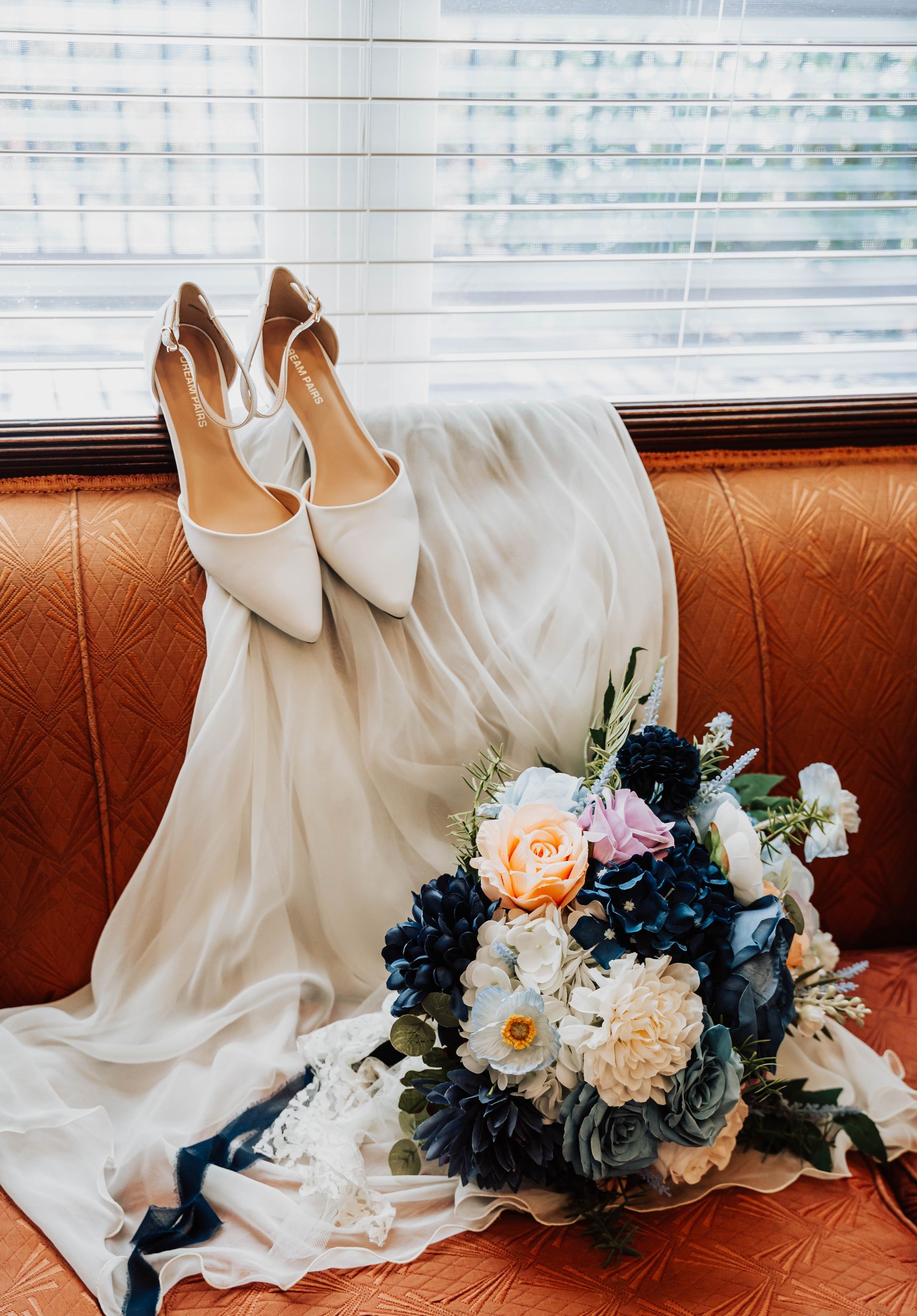 White heels and a flowing dress beside a dark floral bouquet on a rust-colored sofa.