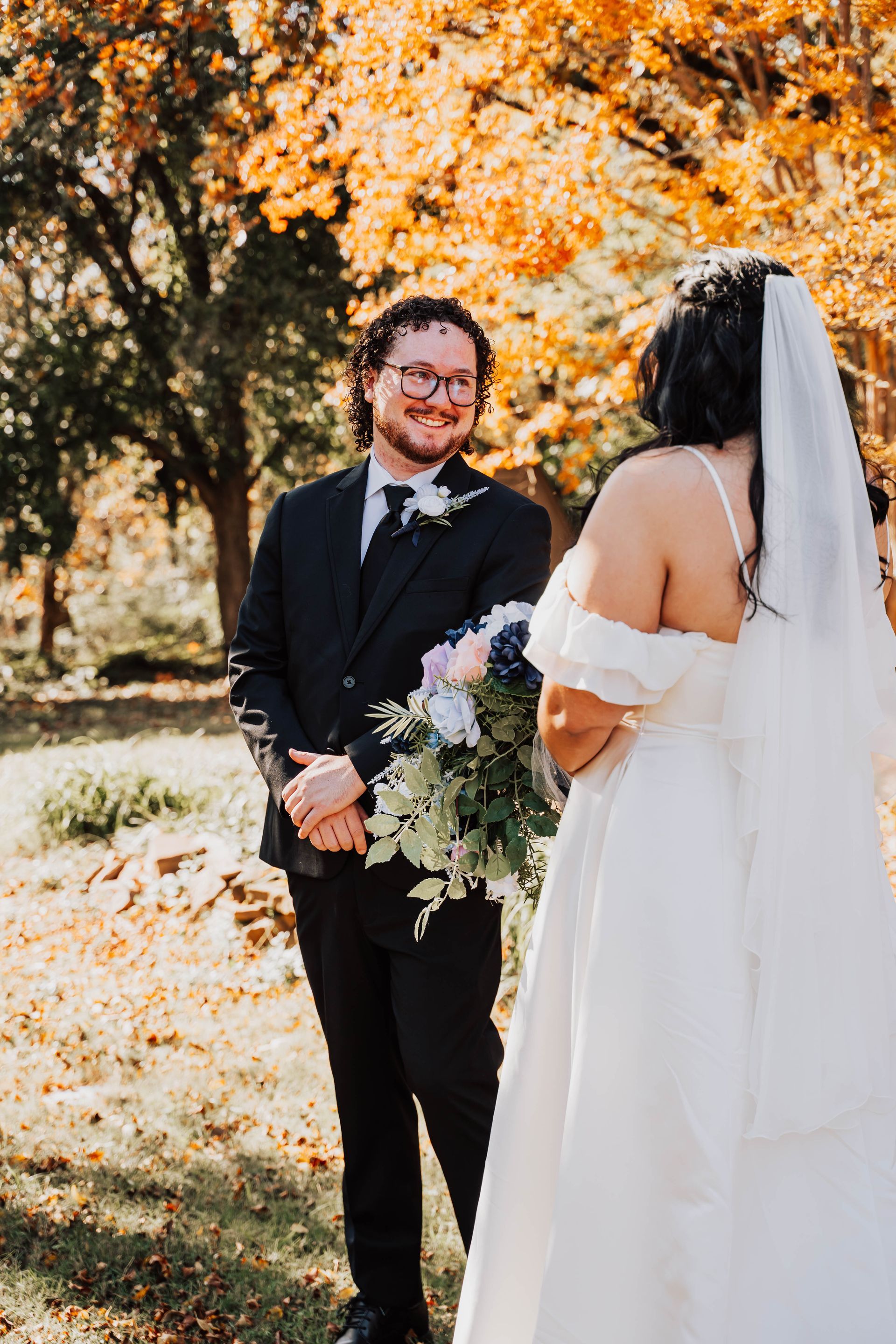 Bride and groom smiling beneath autumn trees, holding a bouquet outdoors.