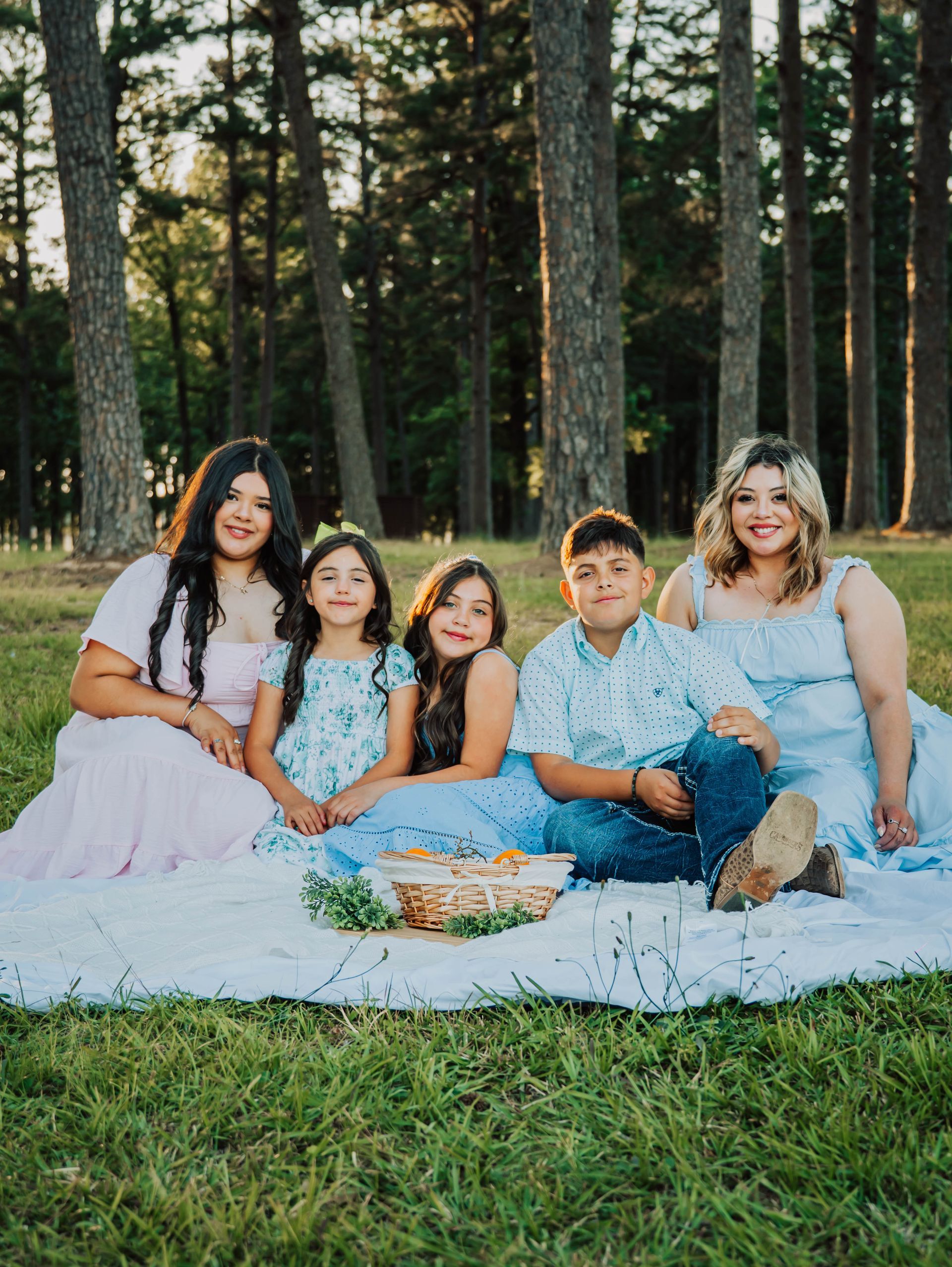 Family sitting on a white blanket in a grassy park, smiling with a picnic basket in front of tall trees