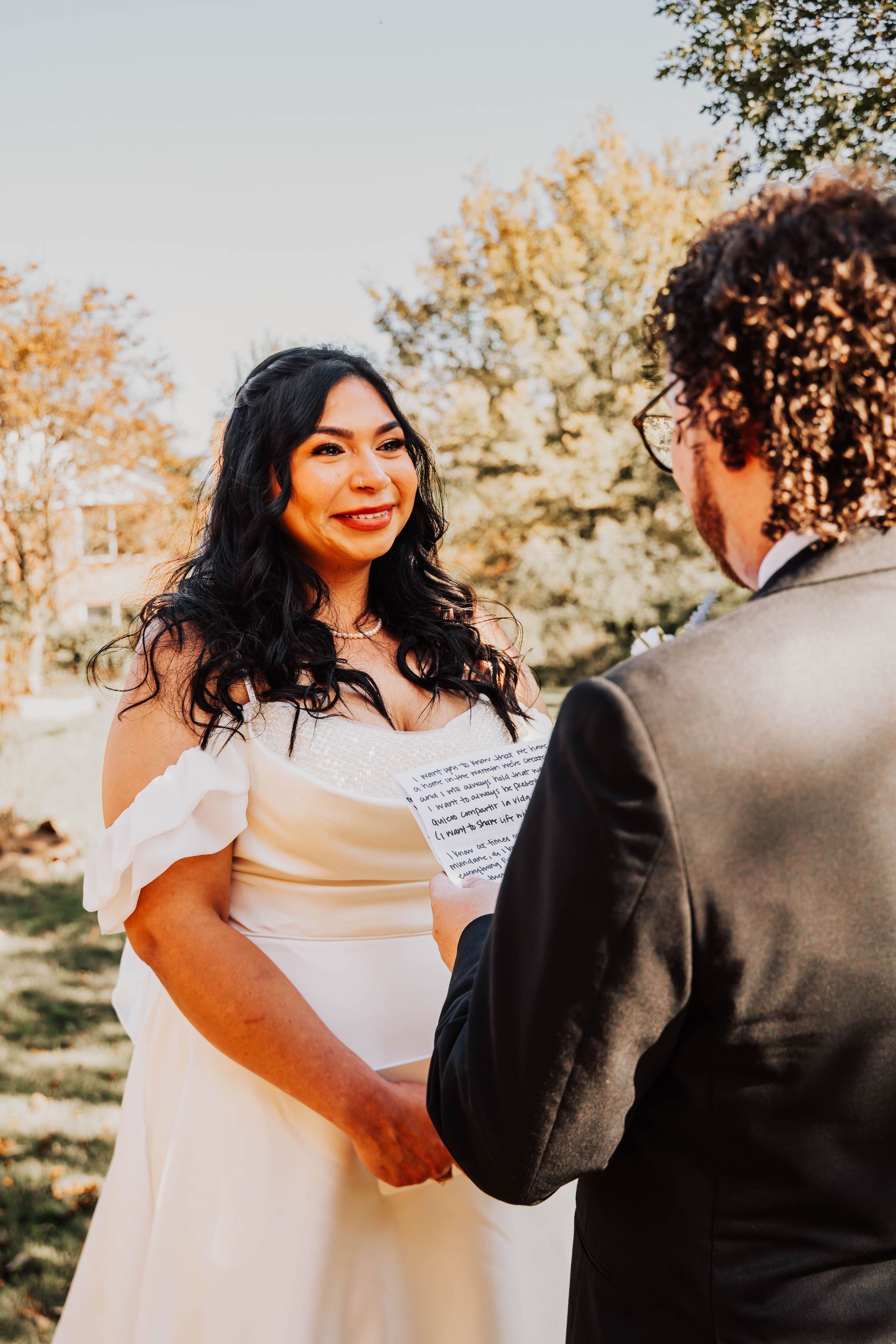 Bride in white dress holding a bouquet, smiling at officiant during outdoor wedding ceremony