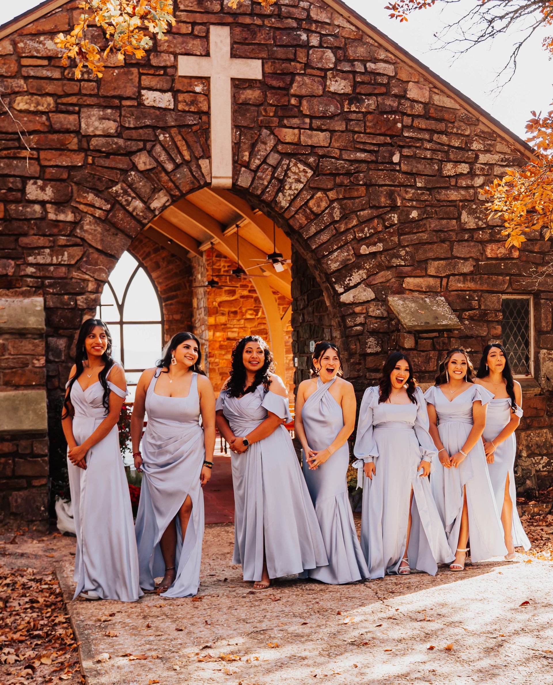 Six women in lavender dresses pose outside a stone chapel with a white cross.