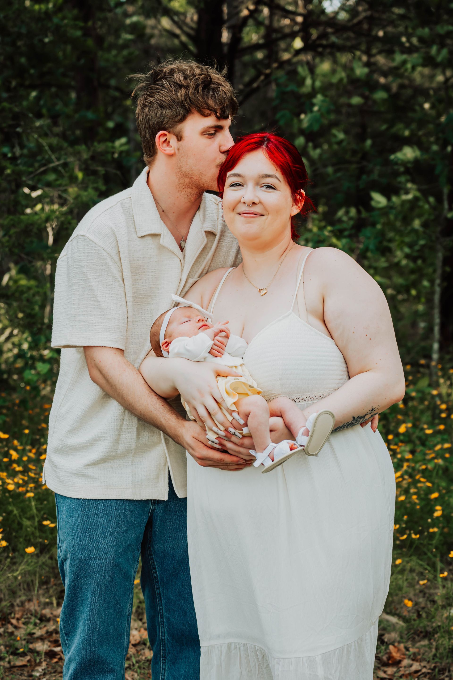 Couple holding a newborn outdoors in a wooded setting, with the man kissing the woman’s forehead.
