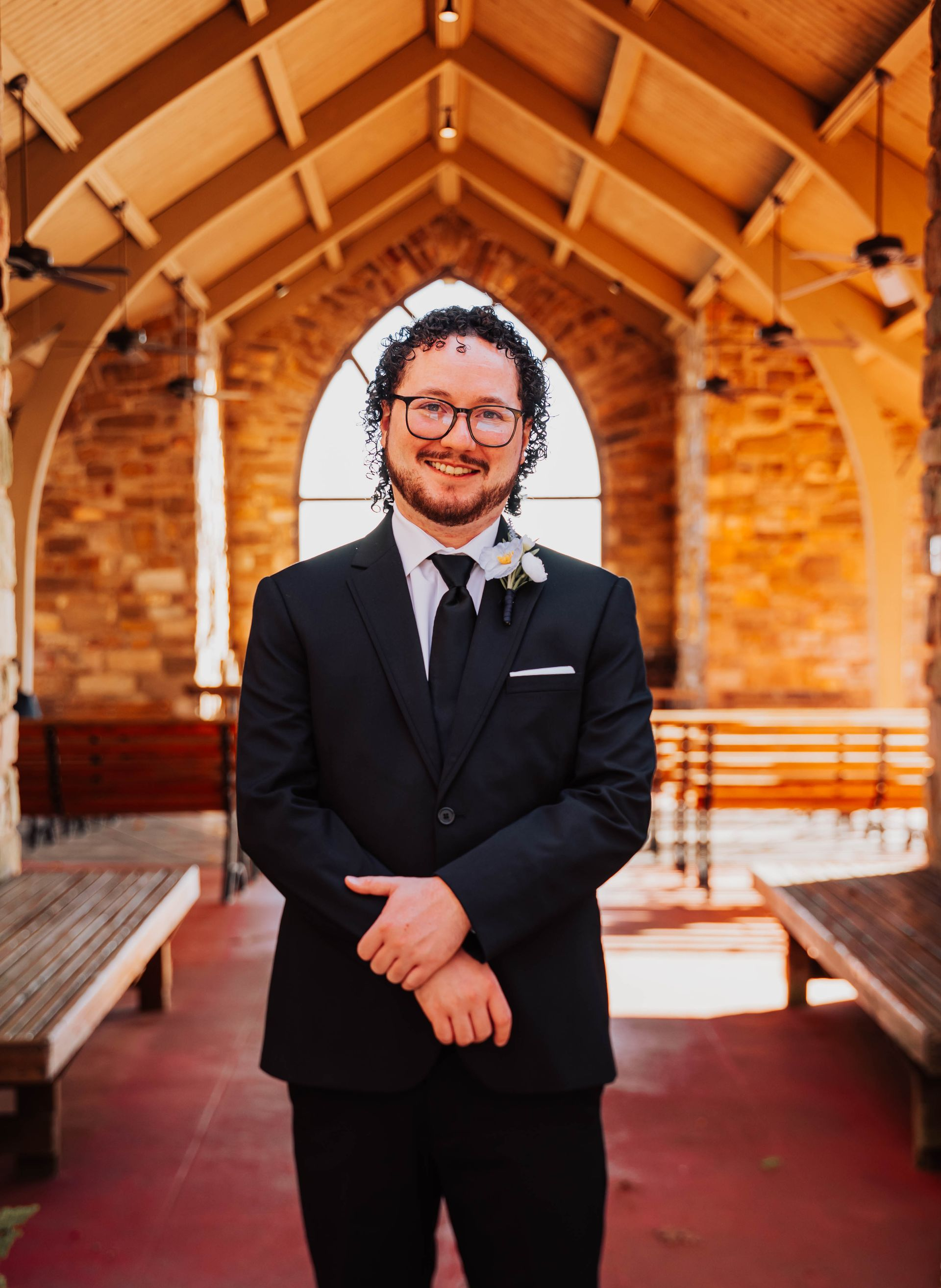 Man in a black suit standing in a wooden chapel aisle, smiling with hands clasped.
