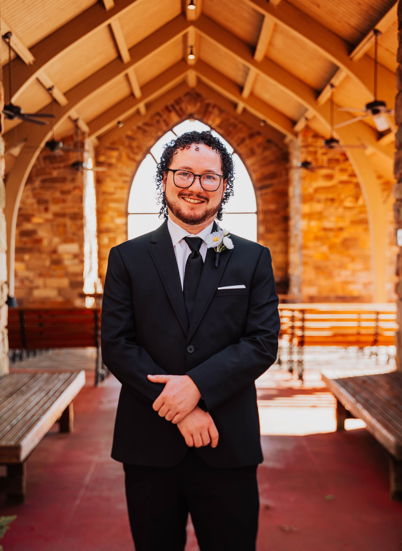 Man in a black suit standing in a wooden chapel aisle, smiling with hands clasped.