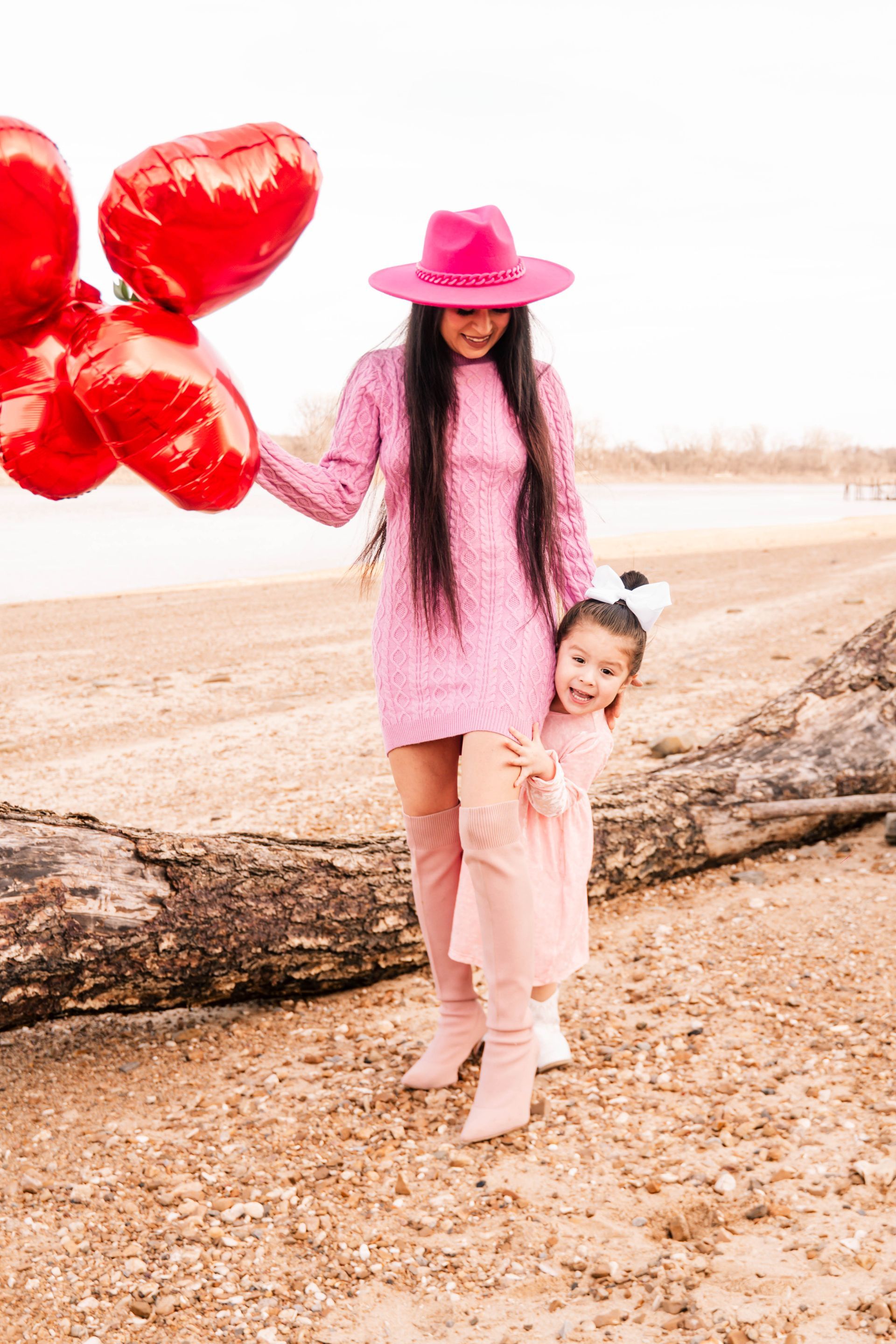 Two children on a beach, one in pink with a hat holding red heart balloons.