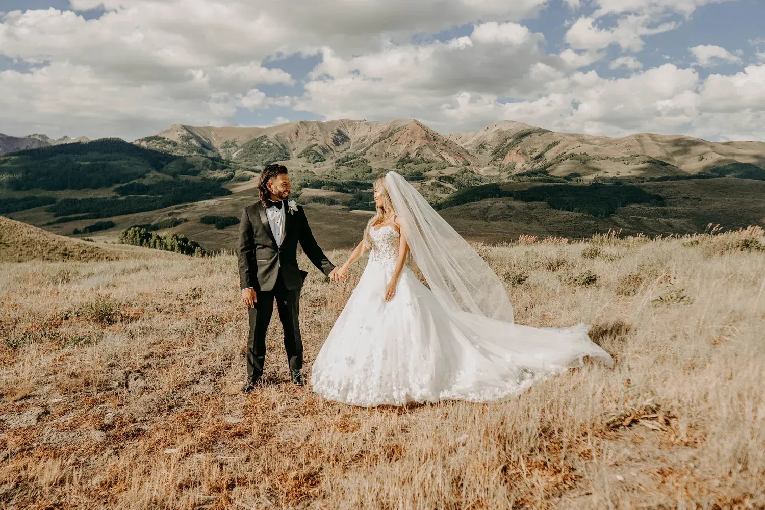 Bride and groom in wedding attire standing in a dry field with mountains and cloudy sky behind them