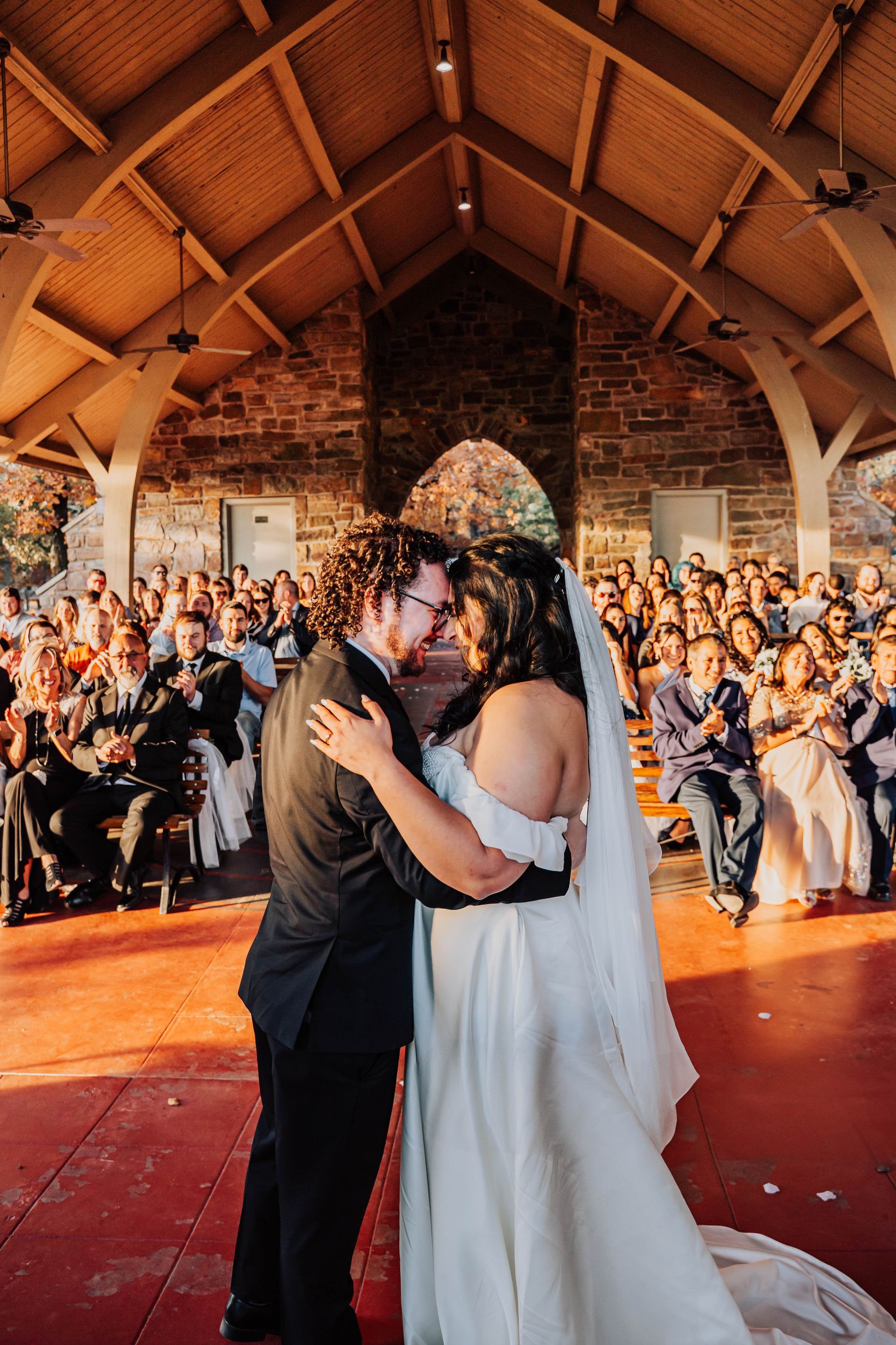 Couple embracing during a wedding dance in a rustic barn, with guests applauding in the background.