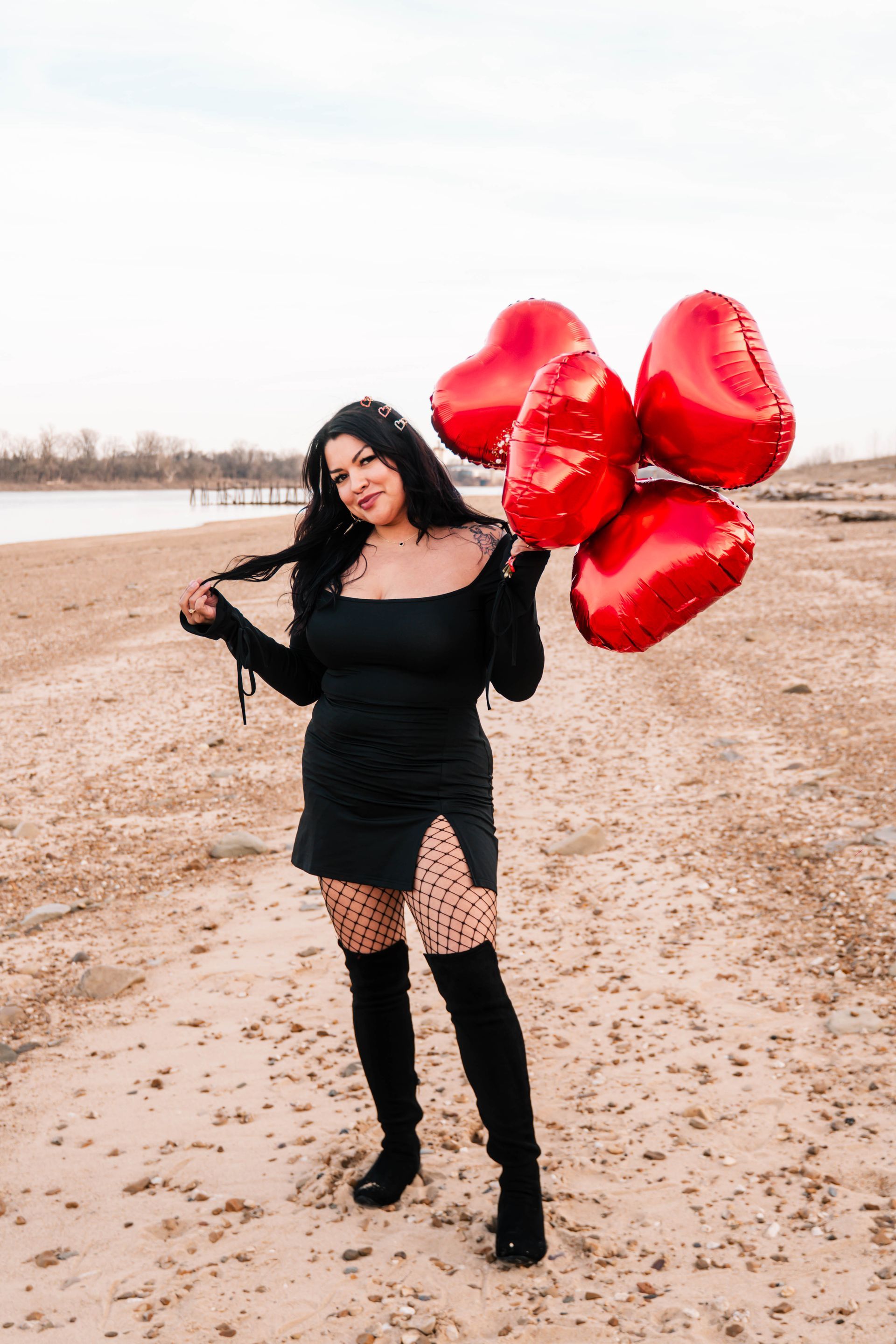 Woman in black dress and boots holding red heart-shaped balloons on a beach