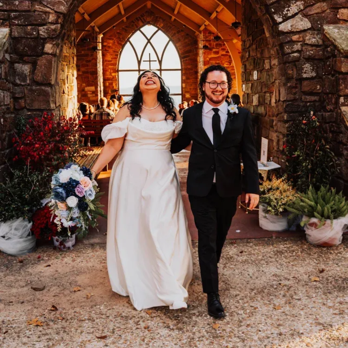 Two people in formal attire walking hand in hand under a stone archway, smiling at an outdoor venue.