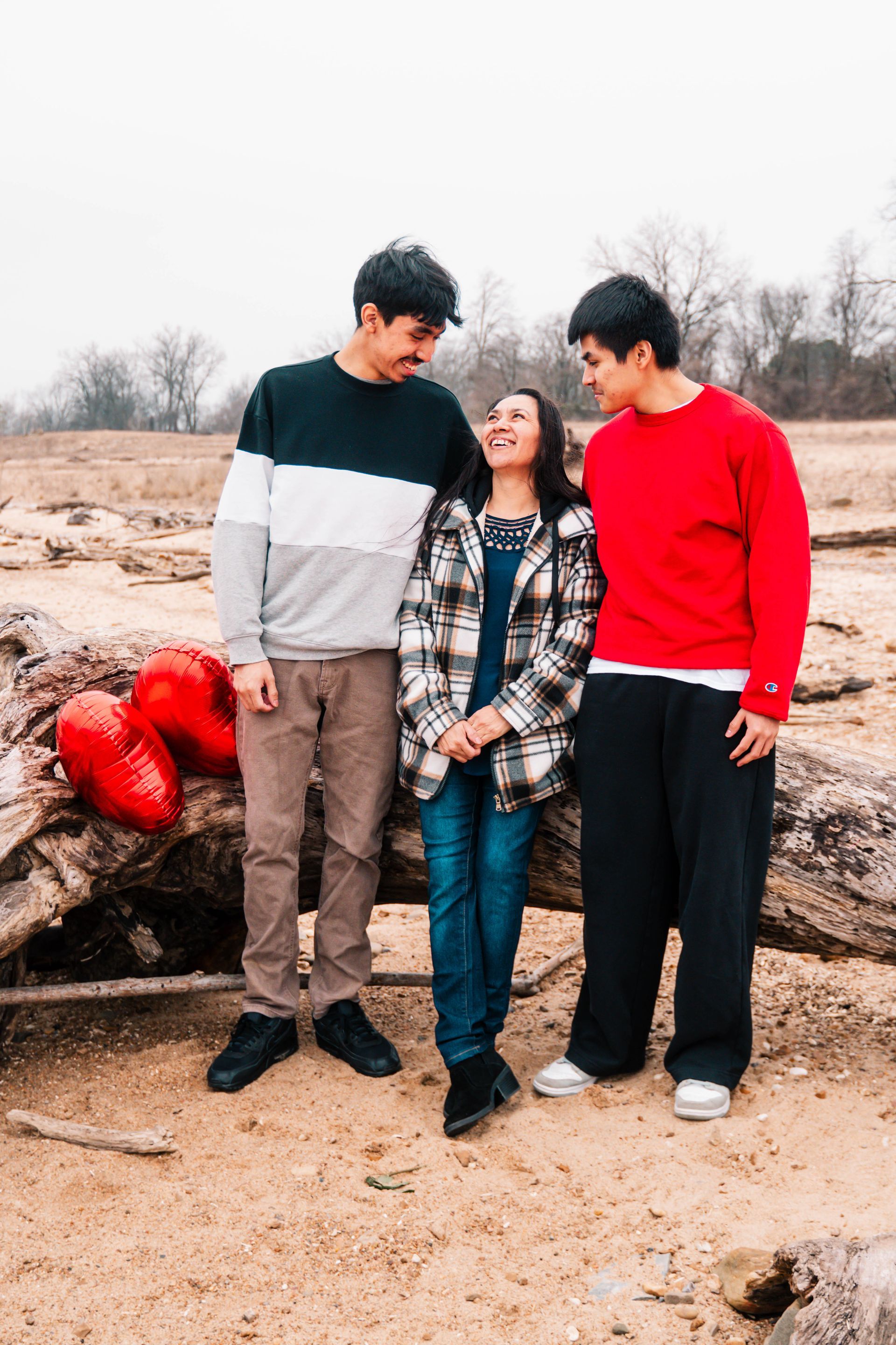 Three people posing outdoors on sandy ground near logs, wearing a black, plaid, and red outfit.