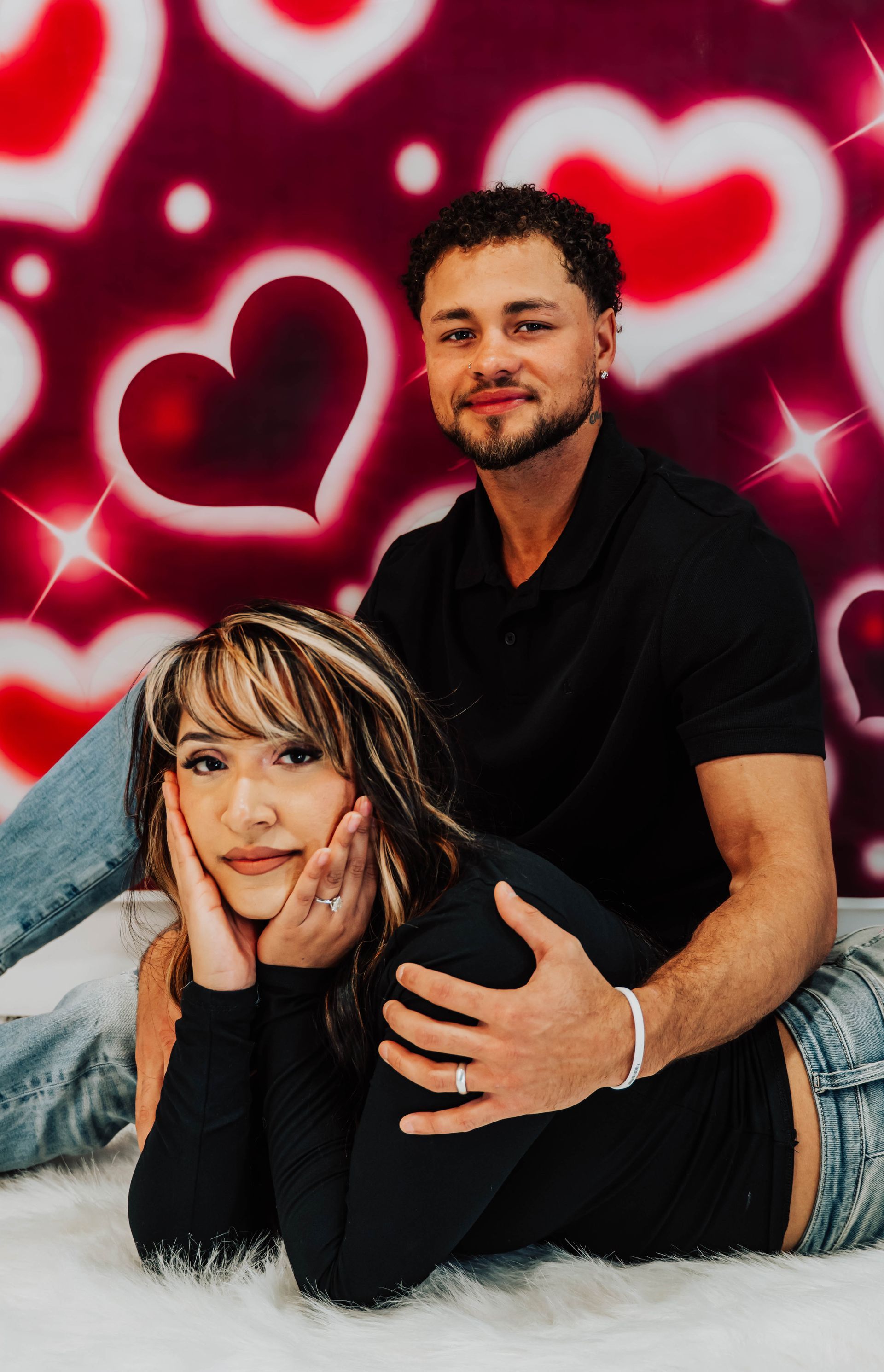 Couple posing together on a white rug in front of a red heart-pattern backdrop