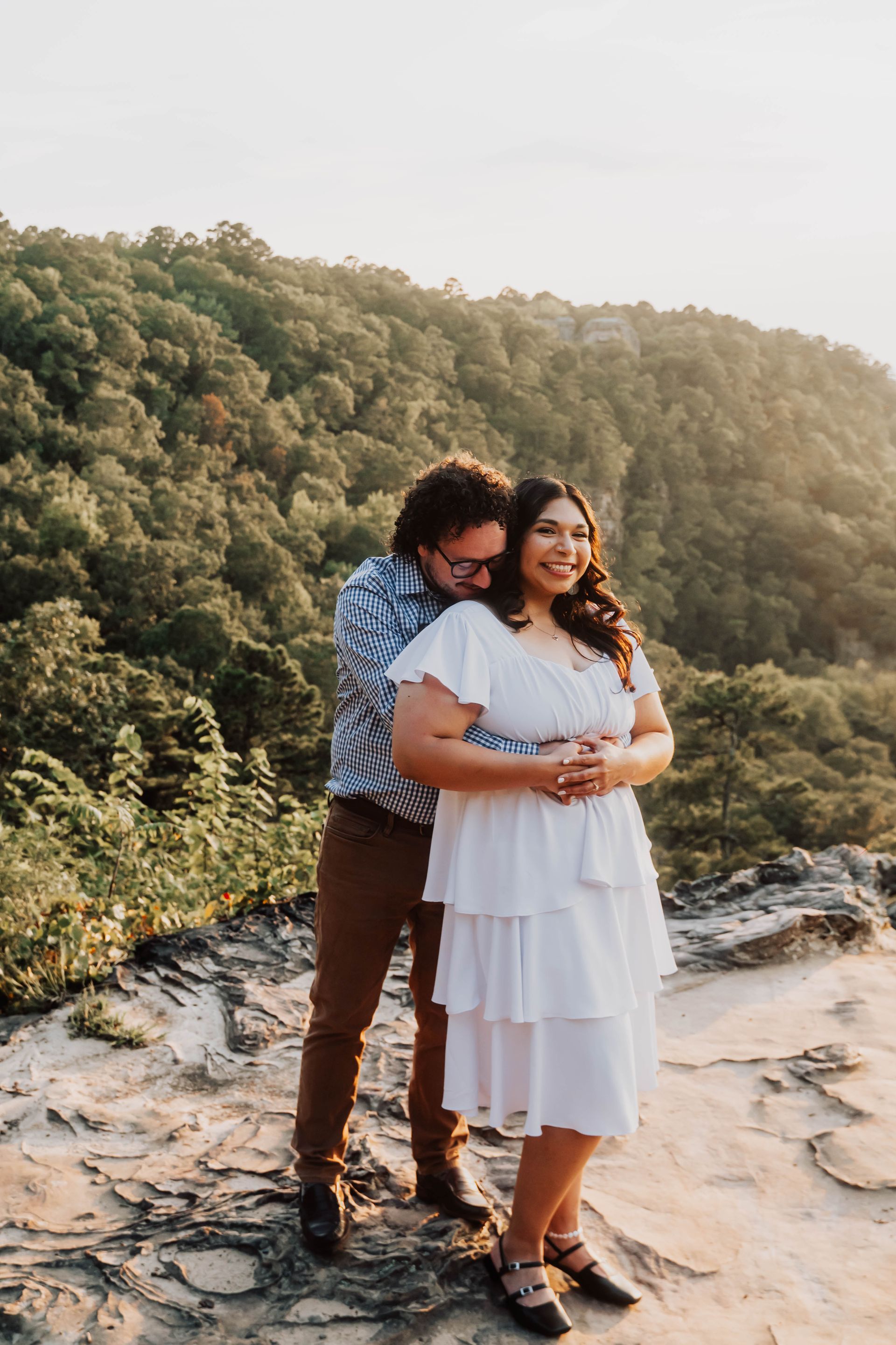 Couple hugging on a rocky overlook with forested hills in the background, in soft golden light