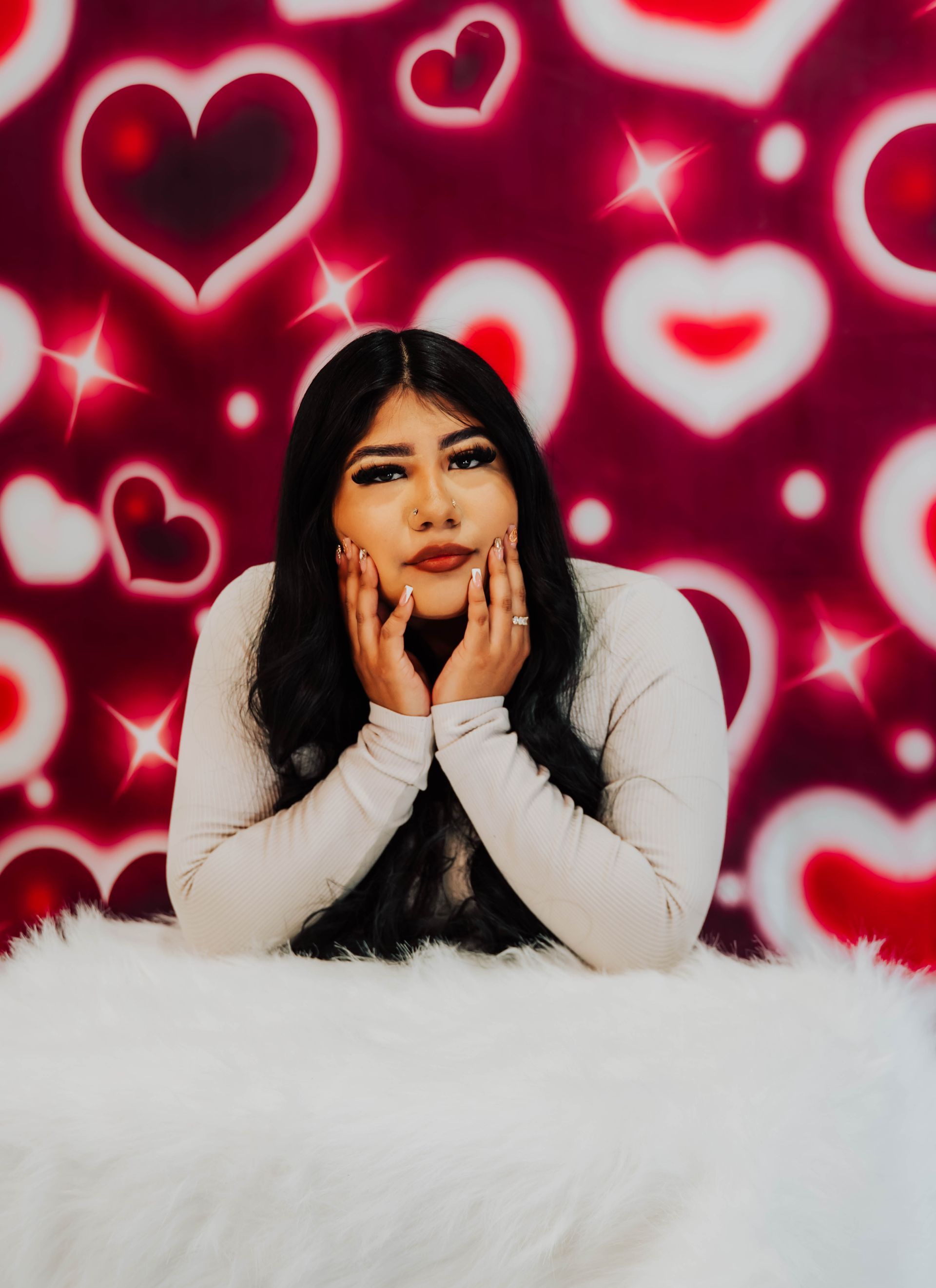 Person posing with hands on cheeks against a red heart-patterned background, wearing white fur.