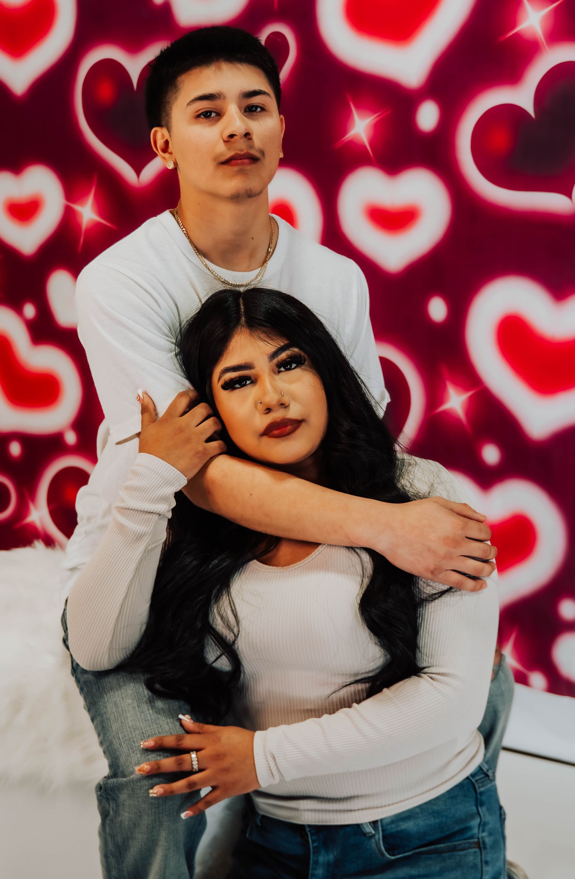 Two people pose in front of a red heart-patterned backdrop, wearing white tops and looking at the camera.