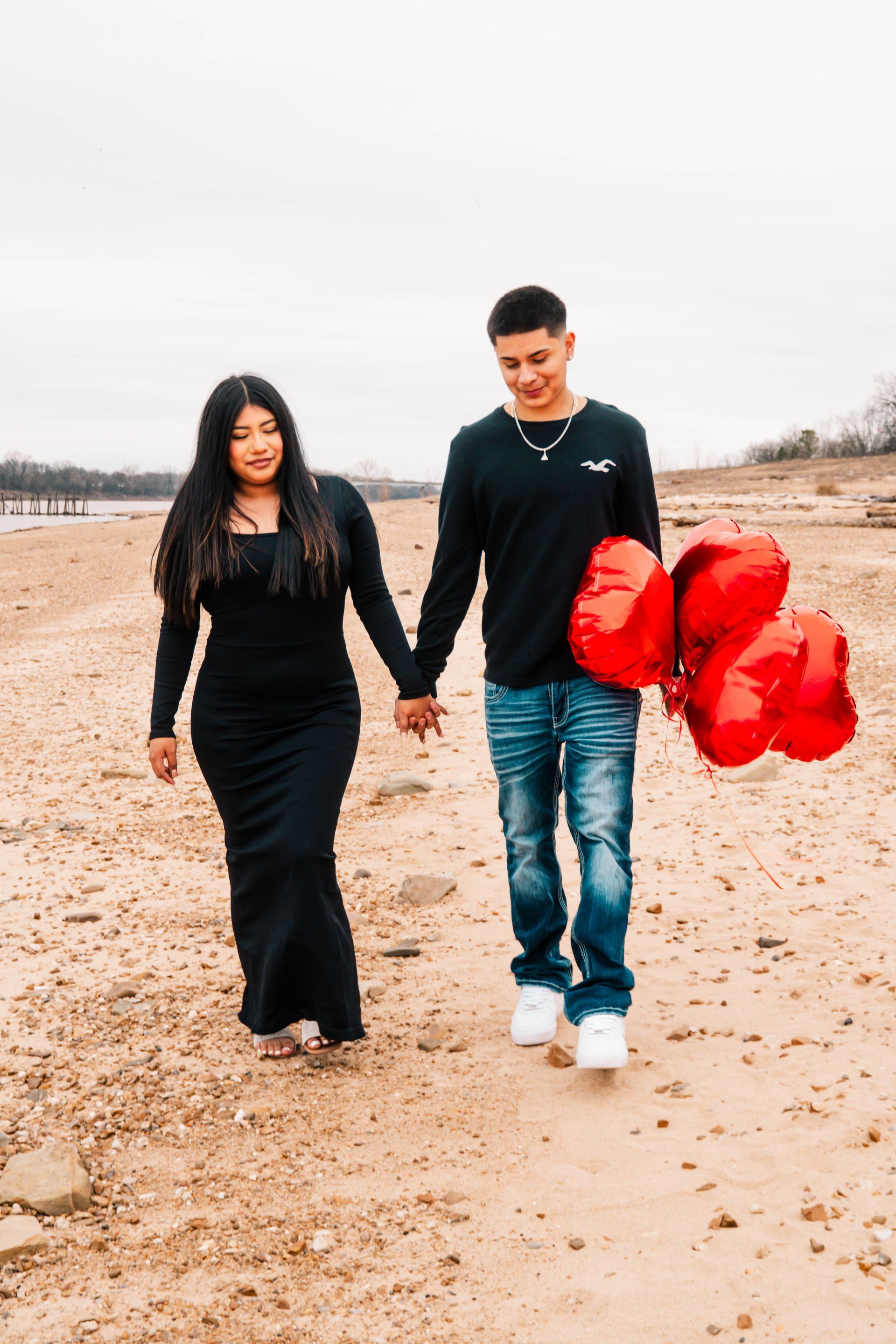 Couple walking hand in hand on a sandy beach, man holding bright red heart balloons